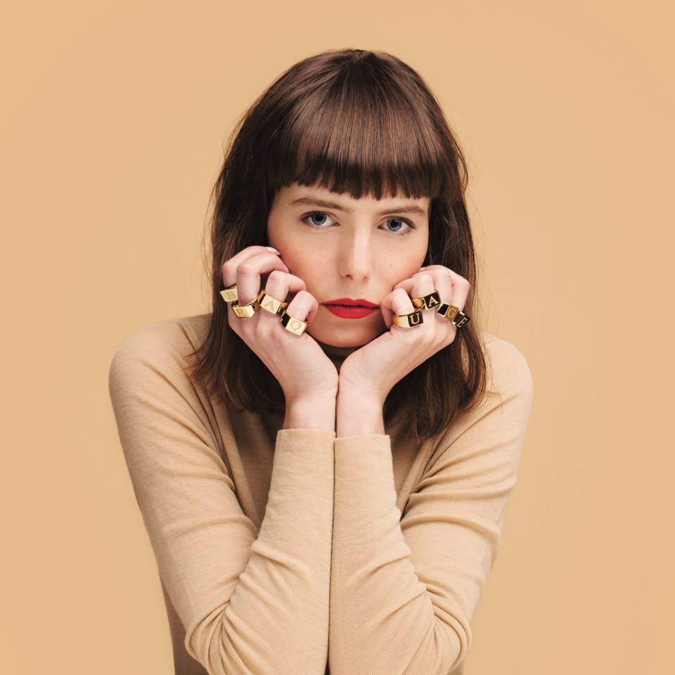 Femme avec des bagues épelant LAURA, rouge à lèvres rouge, fond beige.