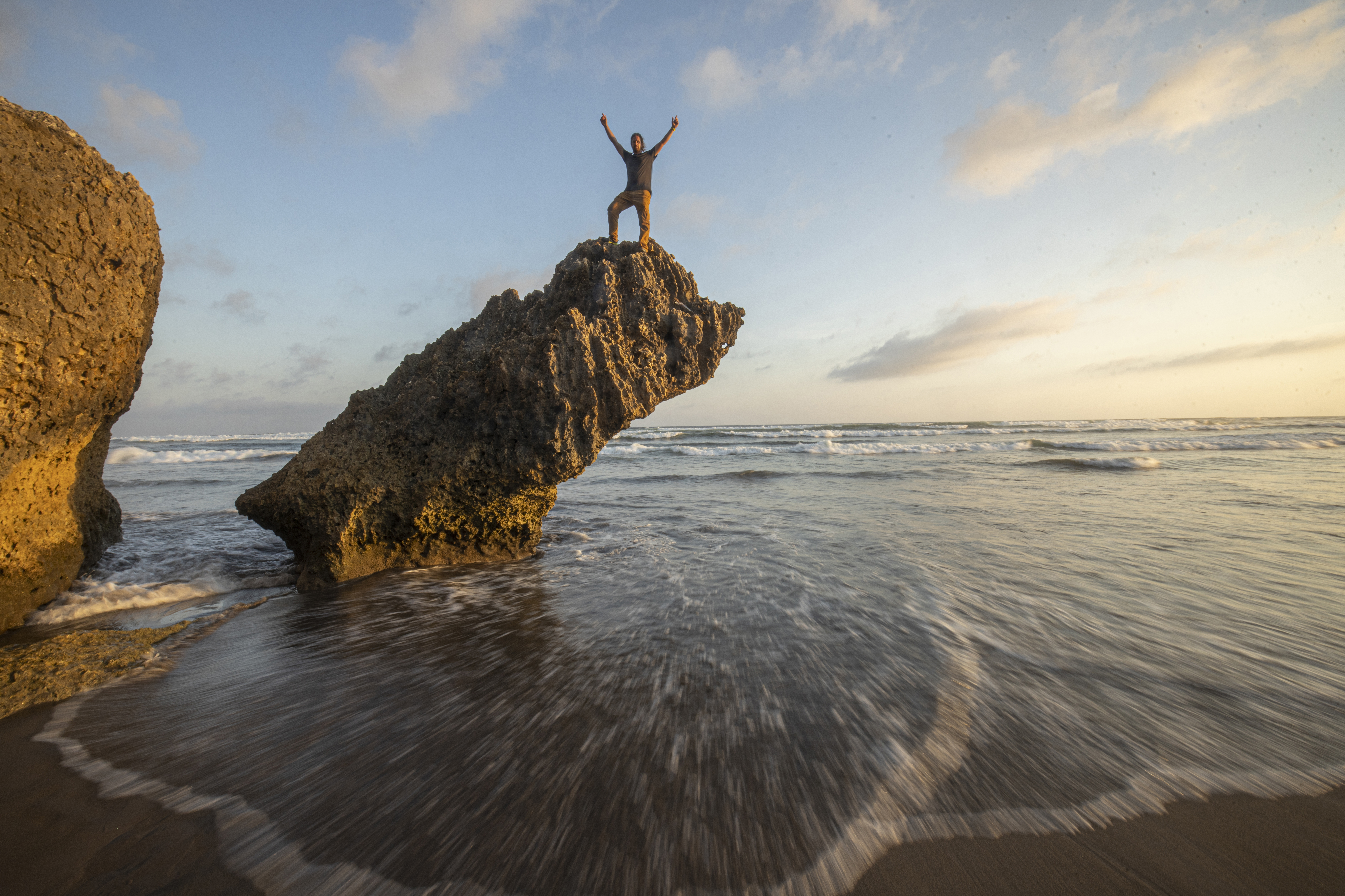 Mann auf Felsen, Arme erhoben, Meerblick, Sonnenuntergang, blauer Himmel, Wasser, Strand.