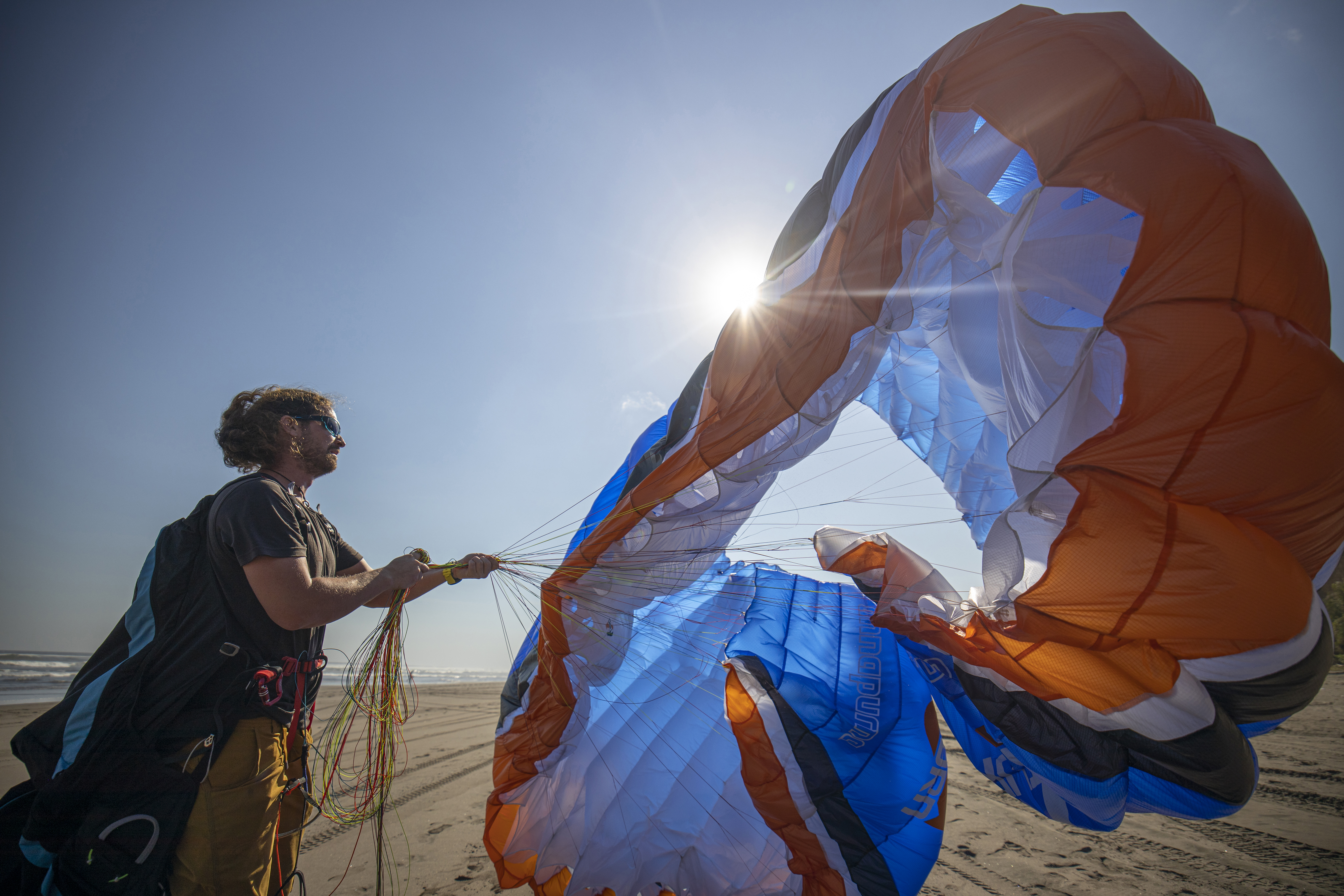 Mann startet bunten Fallschirm am sonnigen Strand. Helle Sonne im Hintergrund.