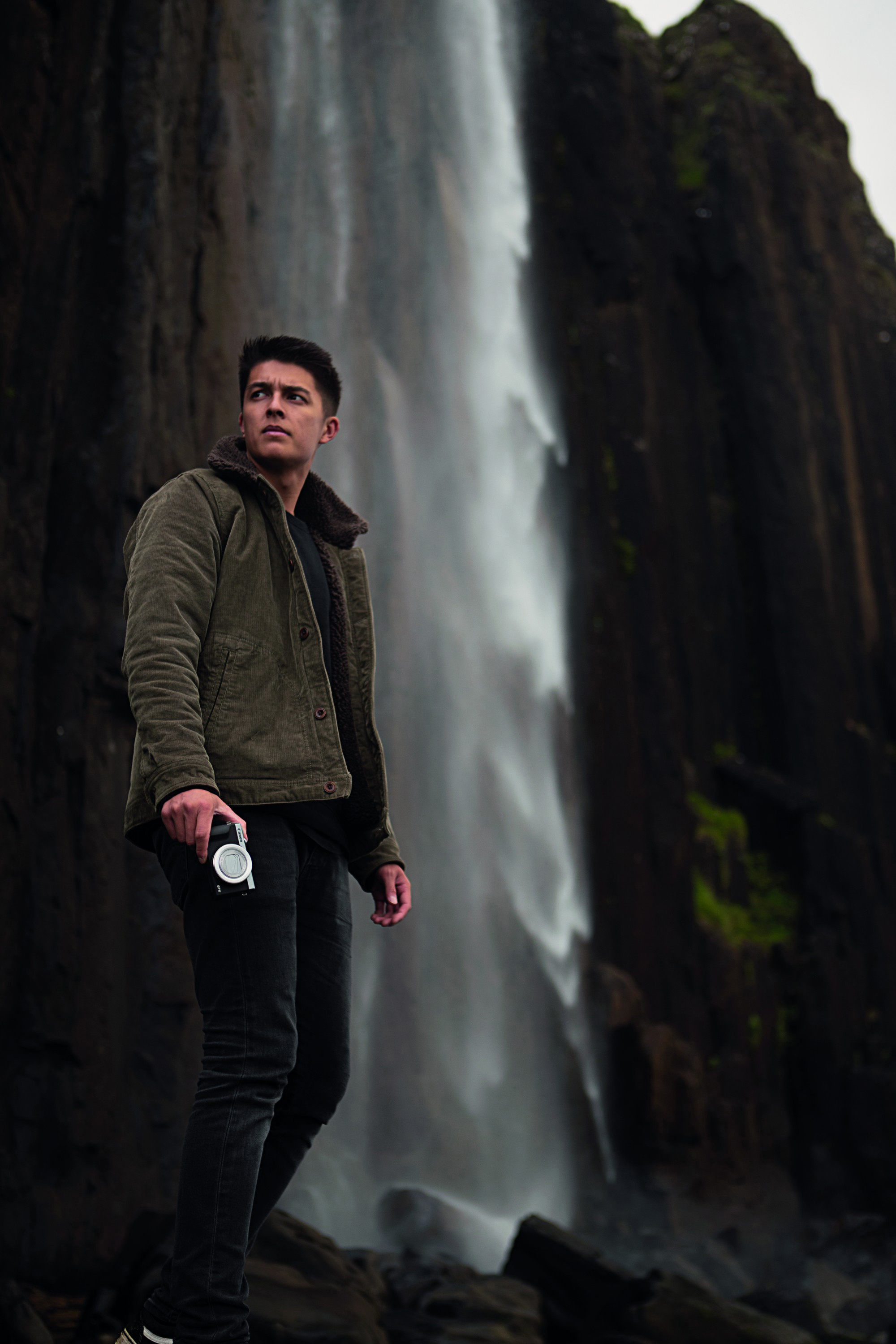 Ein Mann steht vor einem Wasserfall und hält eine Kamera. Er trägt eine Jacke.