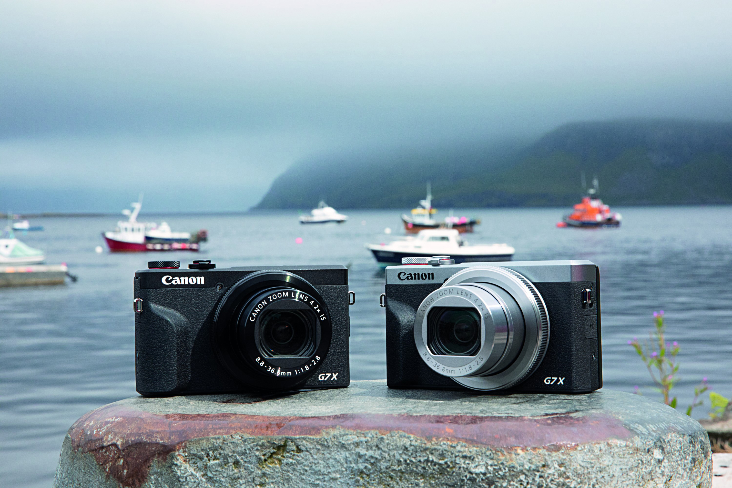 Zwei schwarze und silberne Canon G7X Kameras auf einem Pier mit Booten und Wasser im Hintergrund.