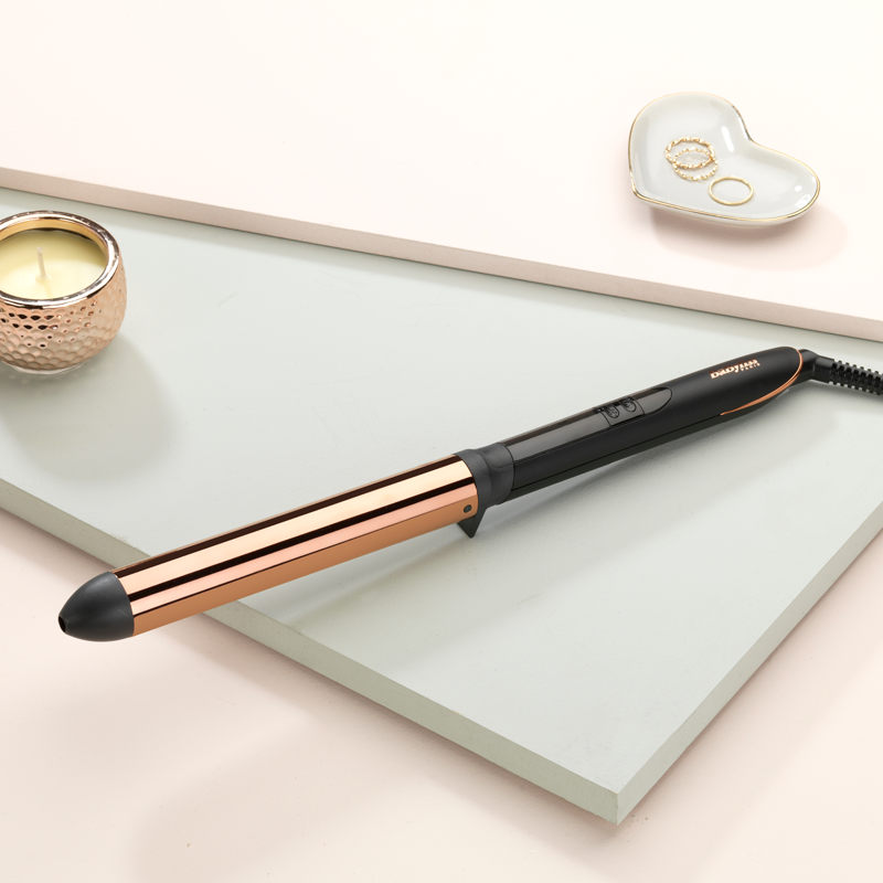 Haarkruller met goud en zwart design, een kaars en een hartvormige schaal met ringen, op een pastel achtergrond.