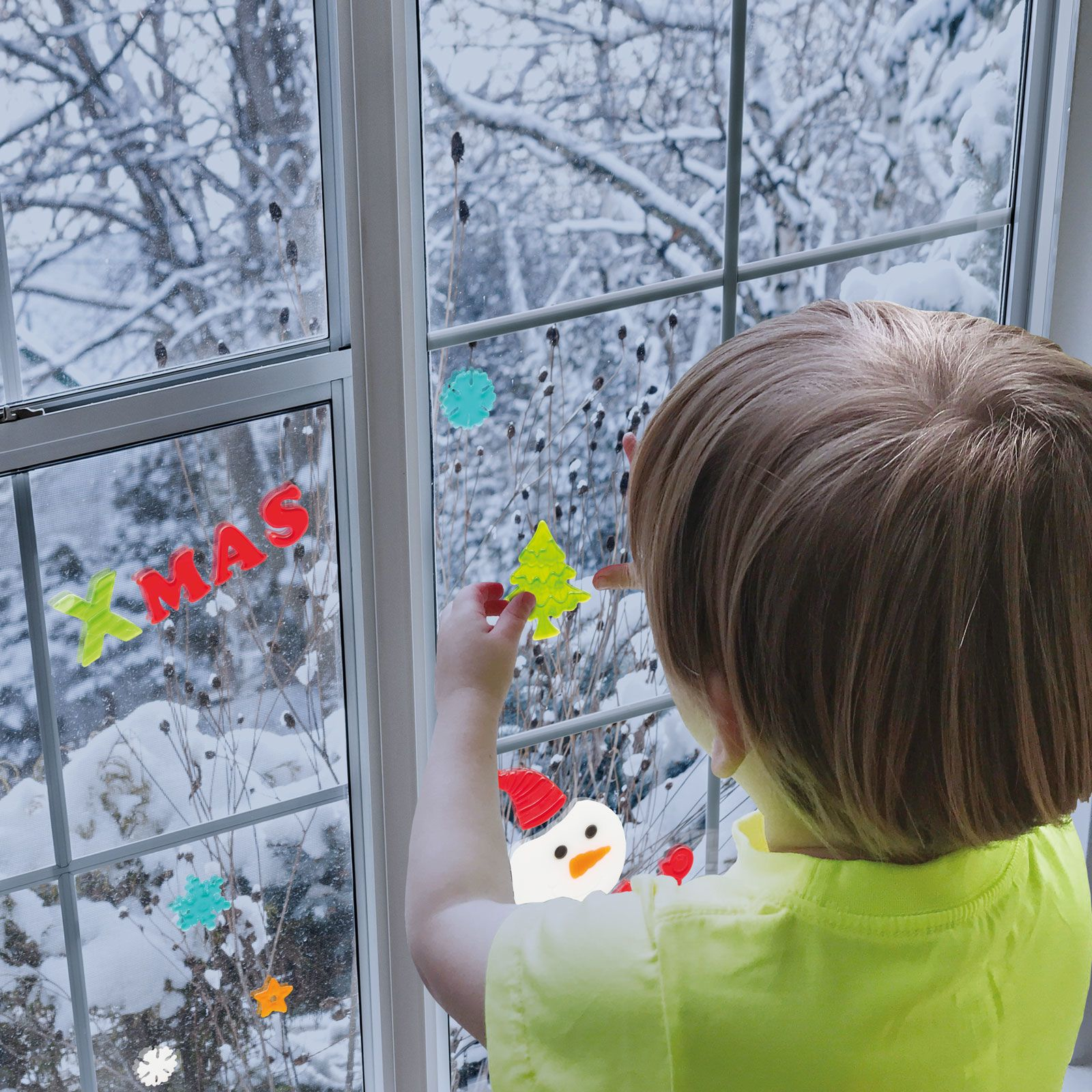 Egy gyermek karácsonyi matricákkal díszít egy ablakot, havas háttérrel az ablakon keresztül.