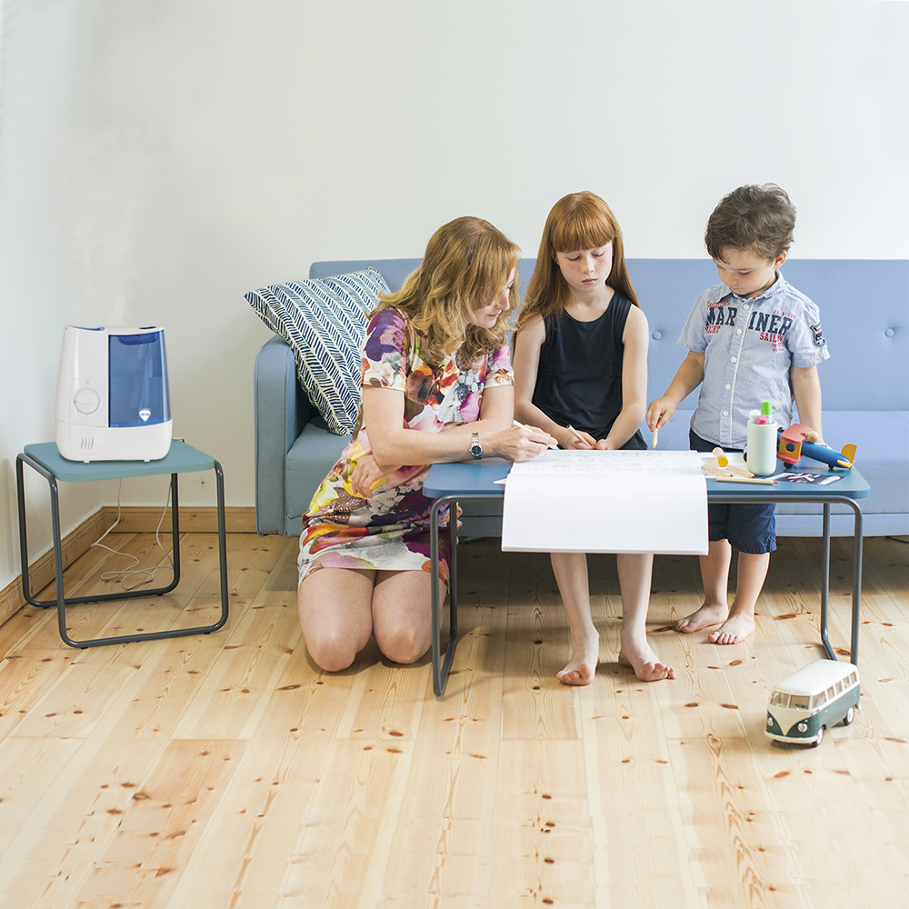 Eine Frau, zwei Kinder und ein Luftbefeuchter sitzen an einem Tisch in einem Raum mit Sofa und Holzboden.