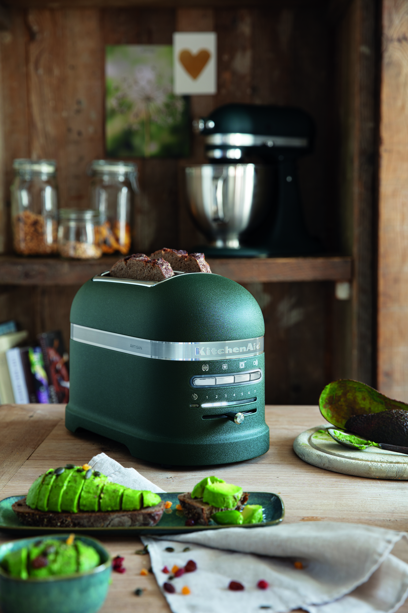 Ein grüner Toaster mit Brotscheiben, Avocado und ein KitchenAid-Mixer auf einem Holztisch.