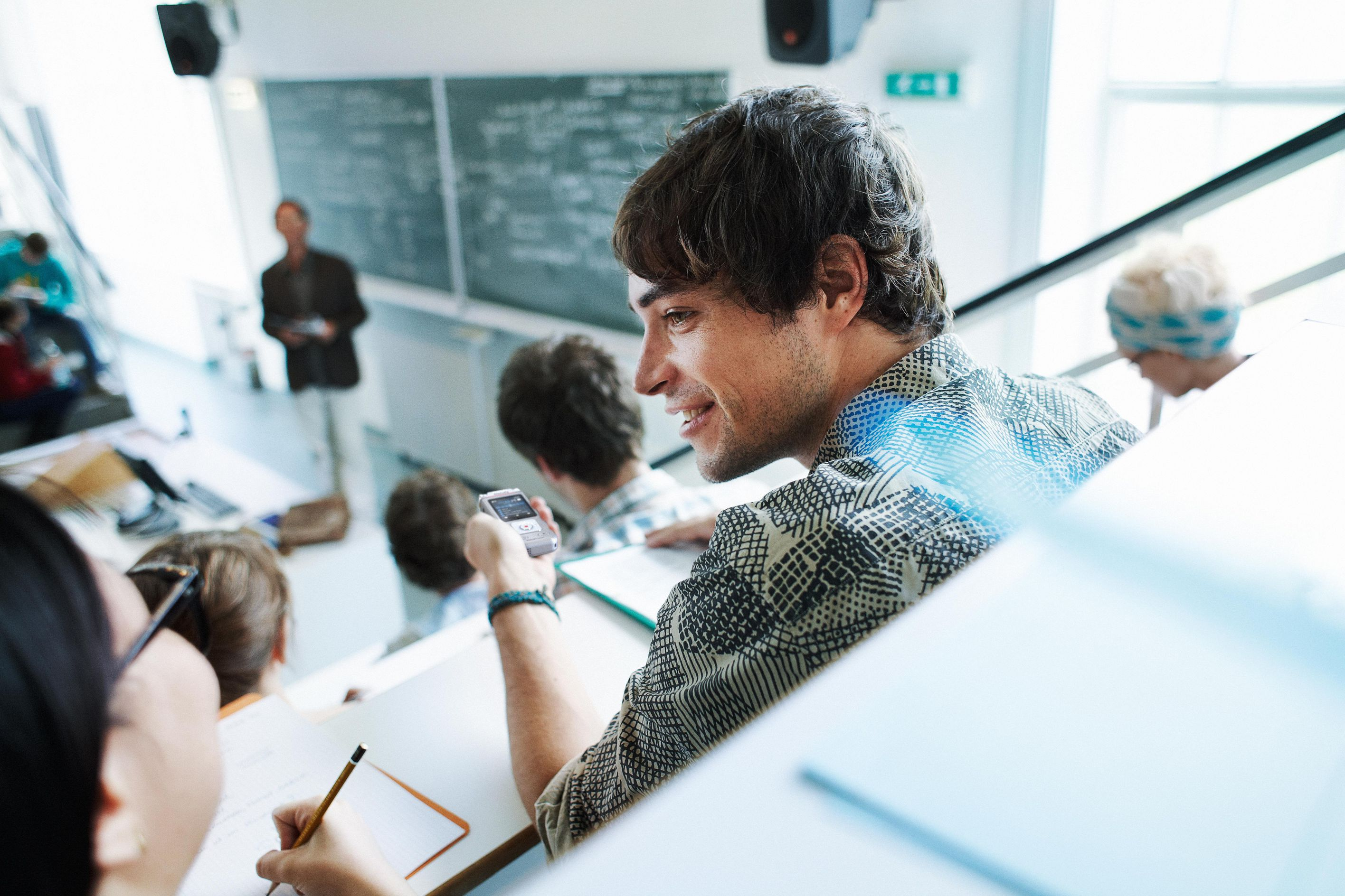 Ein Student im Hörsaal lächelt und hält ein Telefon. Andere Studenten und ein Dozent sind im Hintergrund.