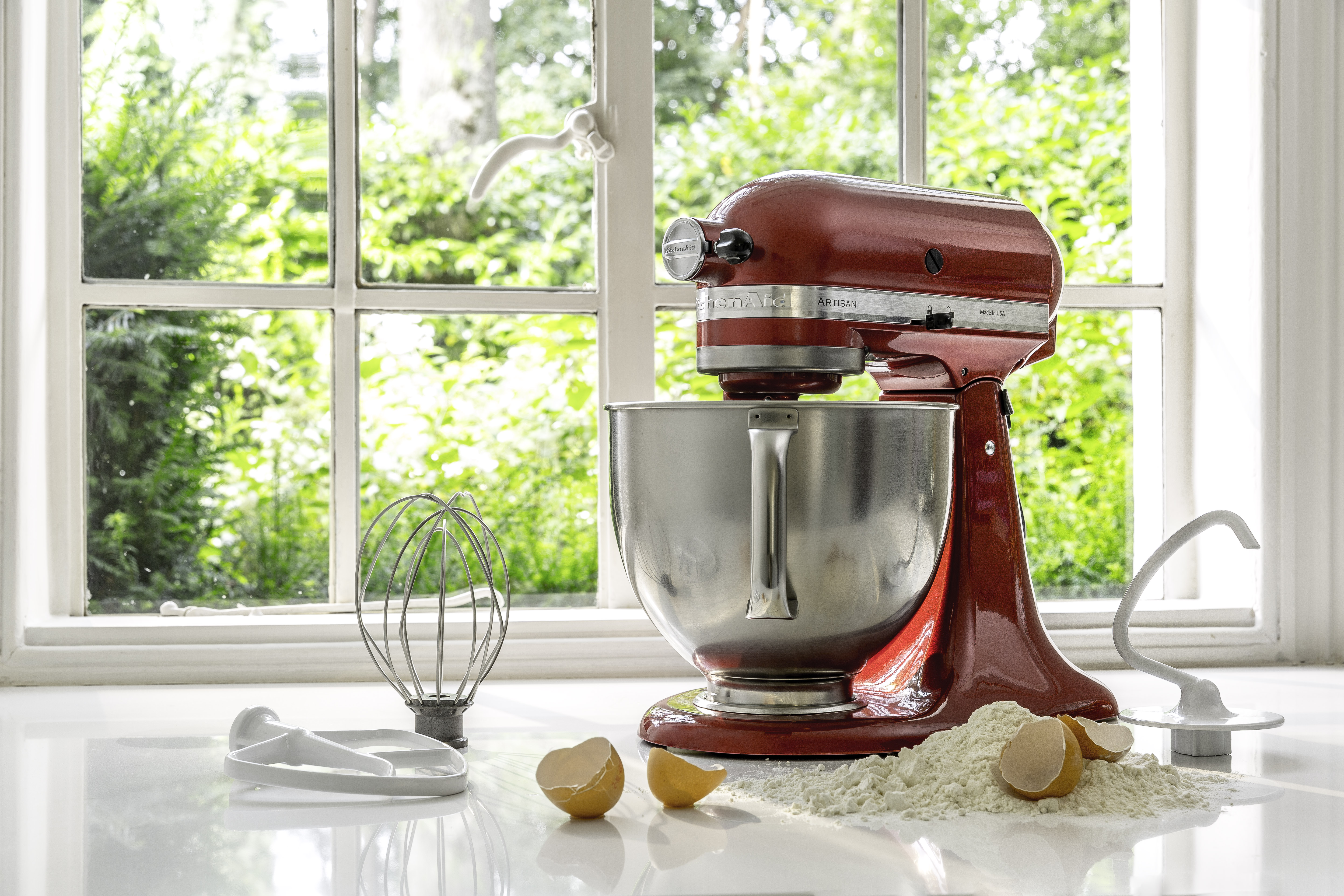 Ein roter KitchenAid-Mixer steht auf einer weißen Oberfläche. Eier und Mehl liegen neben dem Mixer an einem Fenster.