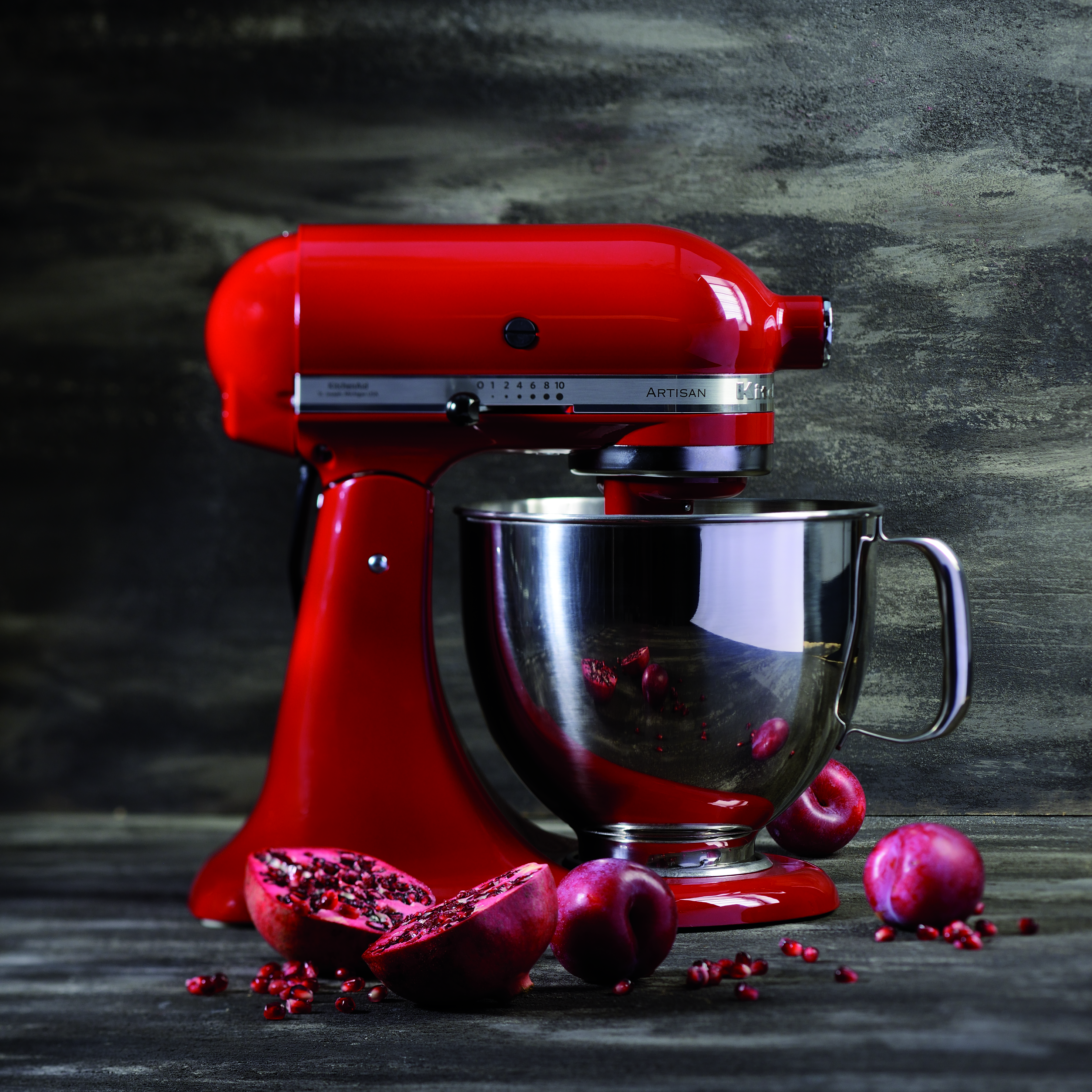Ein roter KitchenAid-Mixer steht neben einer Schüssel, einer halbierten Granatapfel und Pflaumen auf einer Holzoberfläche.