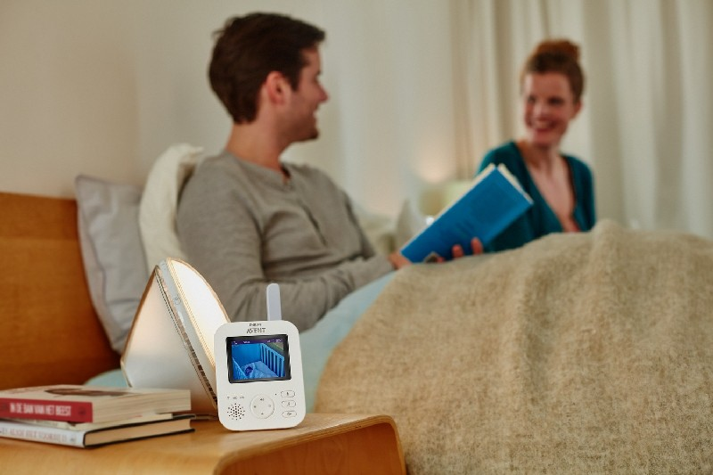 Un vigilabebés en una mesita de noche, con padres en la cama al fondo.