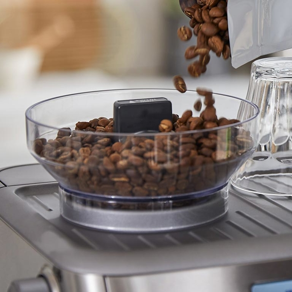 Coffee beans are poured into a clear bowl on top of a coffee machine.