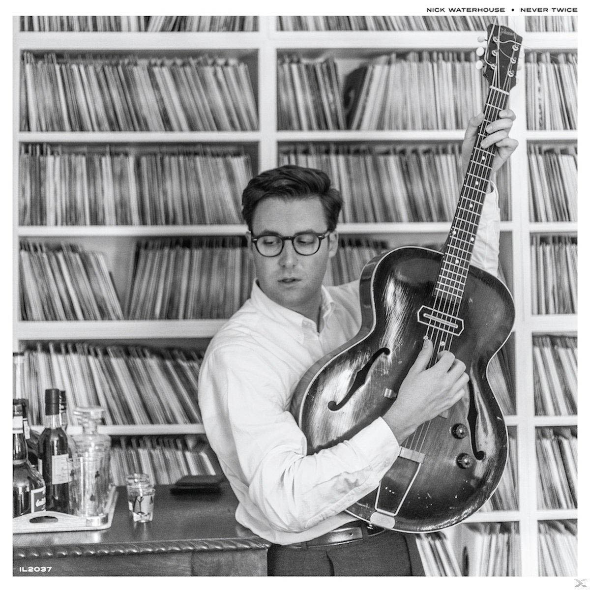 Schwarz-Weiß-Foto eines Mannes mit Brille, der eine Gitarre vor Bücherregalen hält.