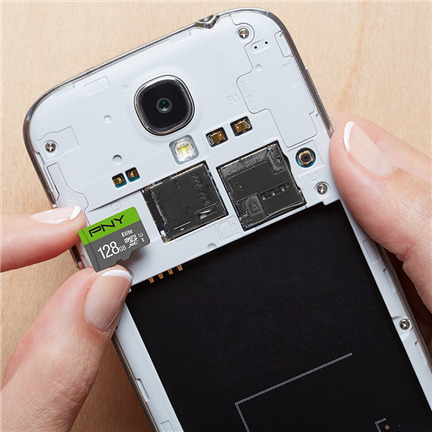 Einsetzen einer PNY 128GB microSD-Karte in die offene Rückseite eines weißen Smartphones auf einem Holztisch.