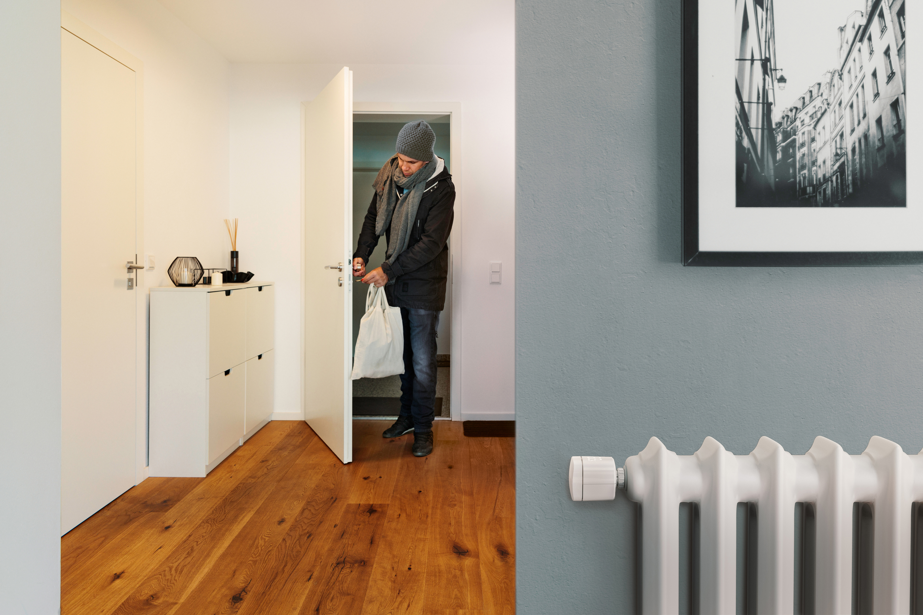 Eine Person öffnet eine weiße Tür. Der Raum hat Holzböden und einen Heizkörper. An der Wand hängt ein Bild.