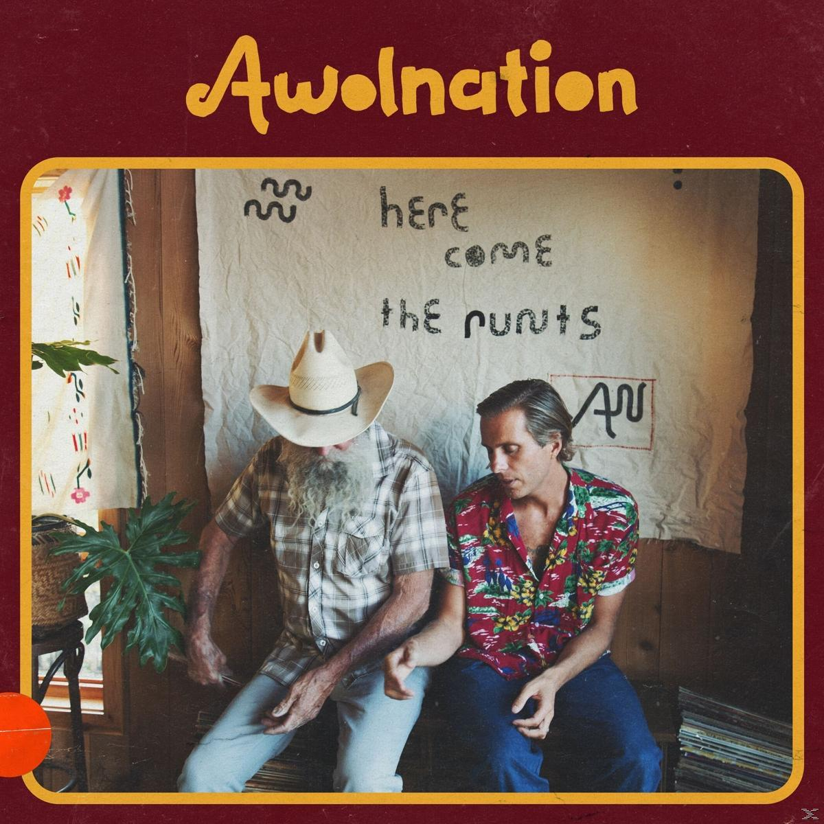 Twee mannen zitten bij een bord met 'Here Come The Runts'. Eén draagt een cowboyhoed en de ander een bloemenhemd.