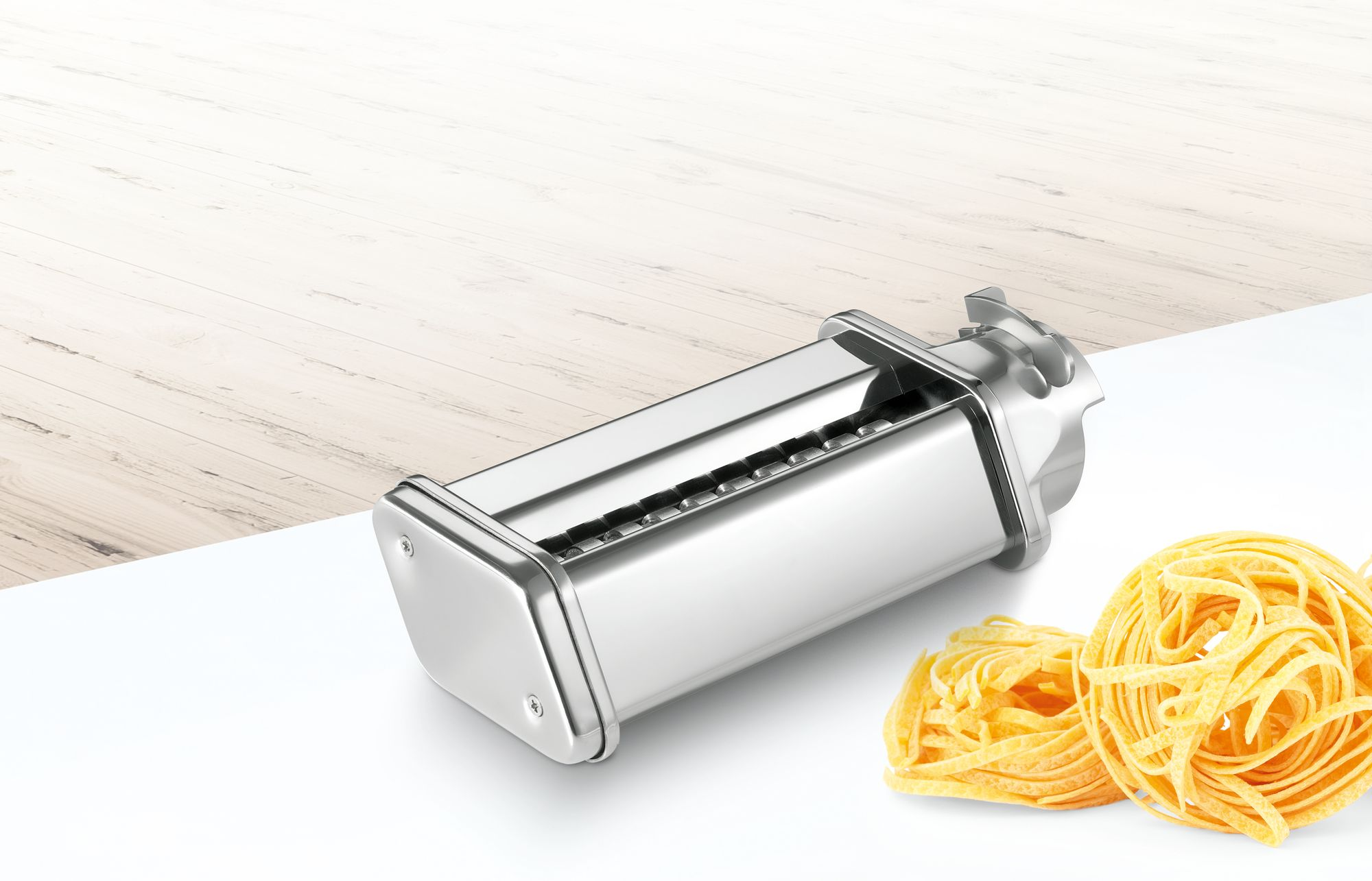 Ezüst tésztagép fettuccine tésztával, fehér felületen, fa háttérrel.