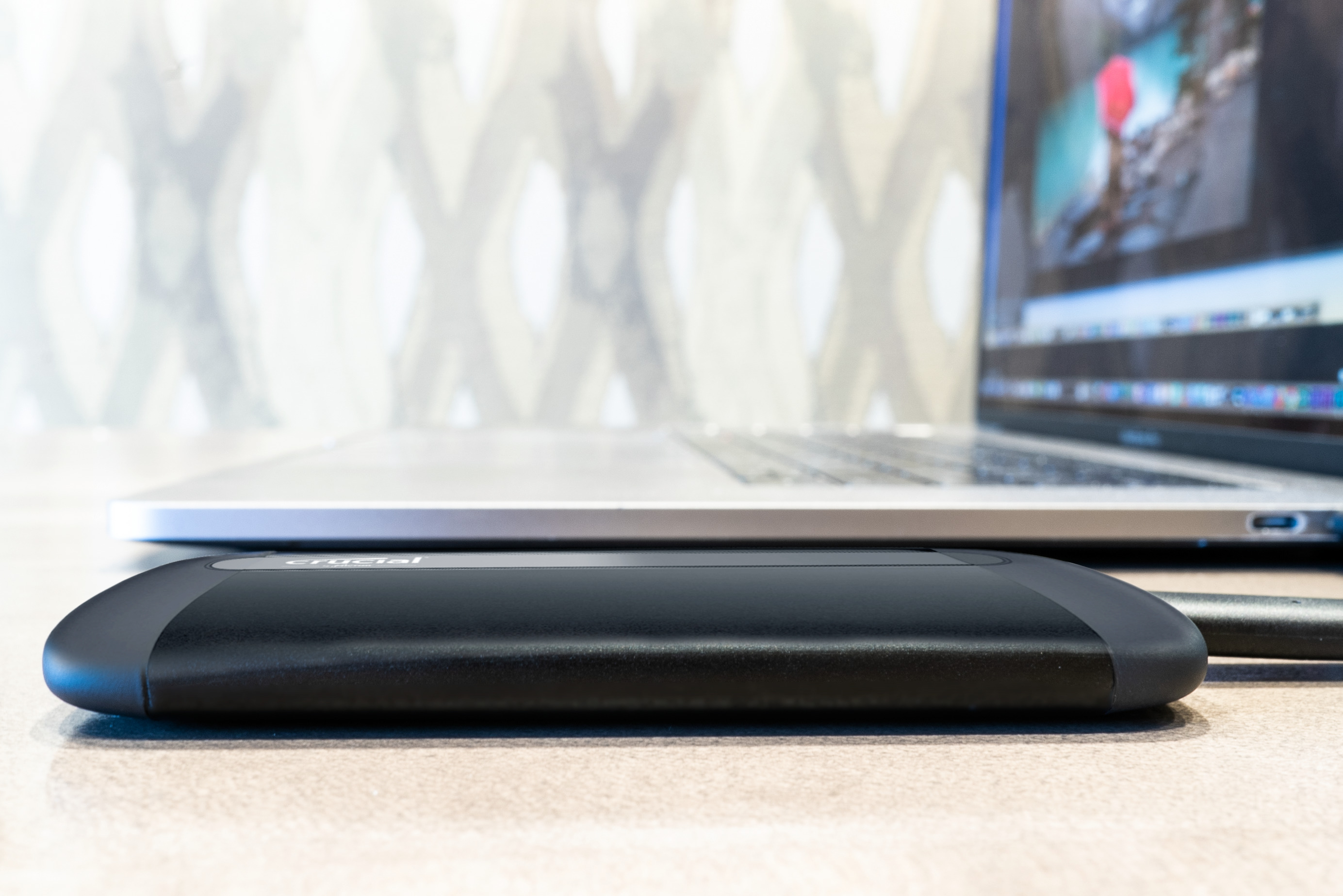 Eine schwarze externe Festplatte liegt auf einer hellen Oberfläche, ein Laptop im Hintergrund. Die Festplatte hat 'crucial'.