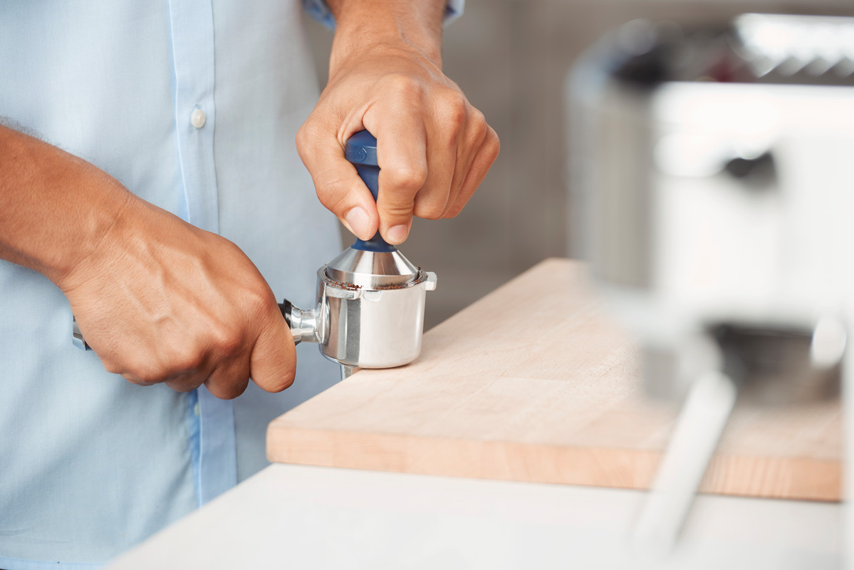 Hände pressen Kaffeepulver in einen Siebträger. Ein Holzbrett und eine Kaffeemaschine sind sichtbar.