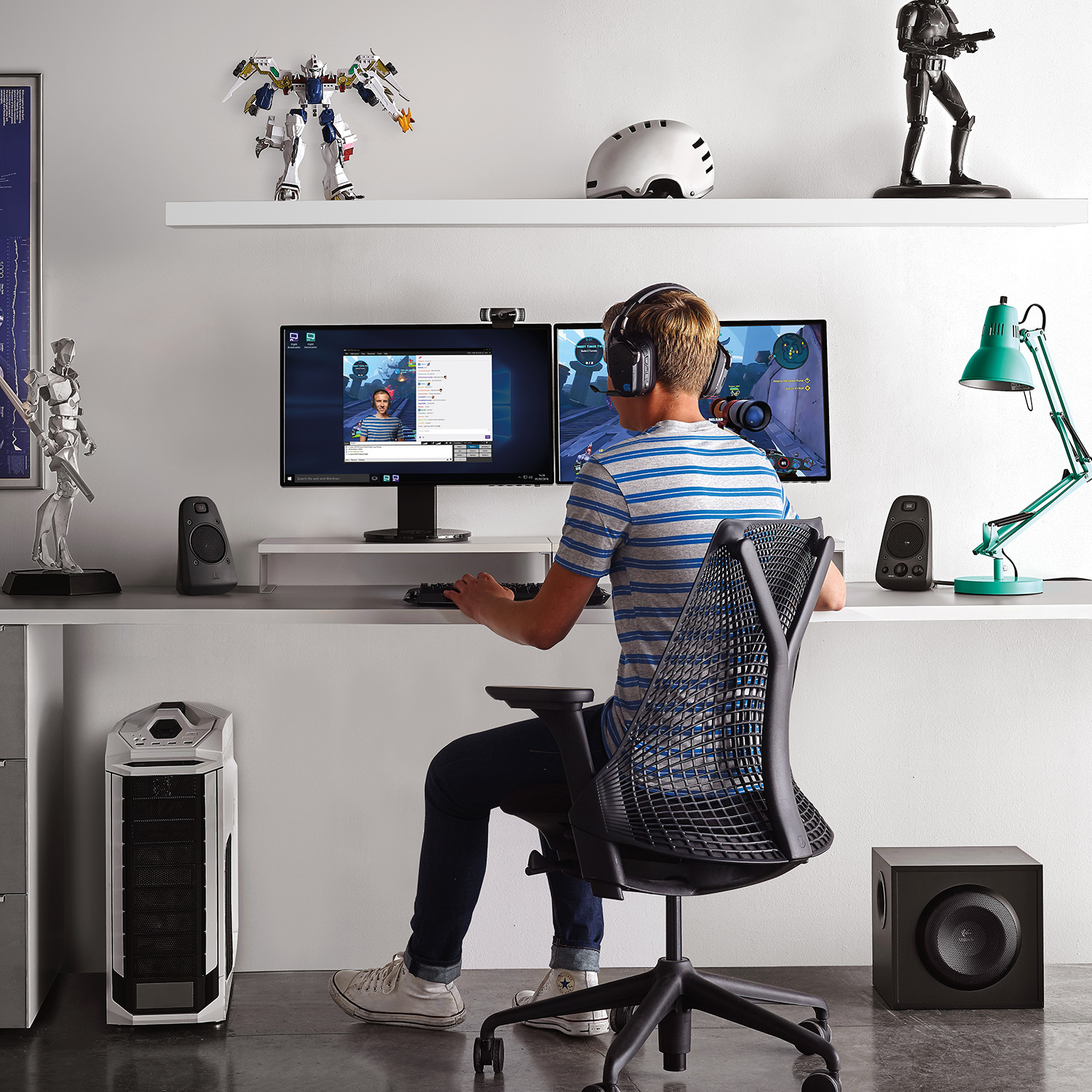 Ein Mann spielt. Er sitzt an einem Schreibtisch mit zwei Monitoren, einem Computer und Lautsprechern. Spielzeug auf einem Regal.