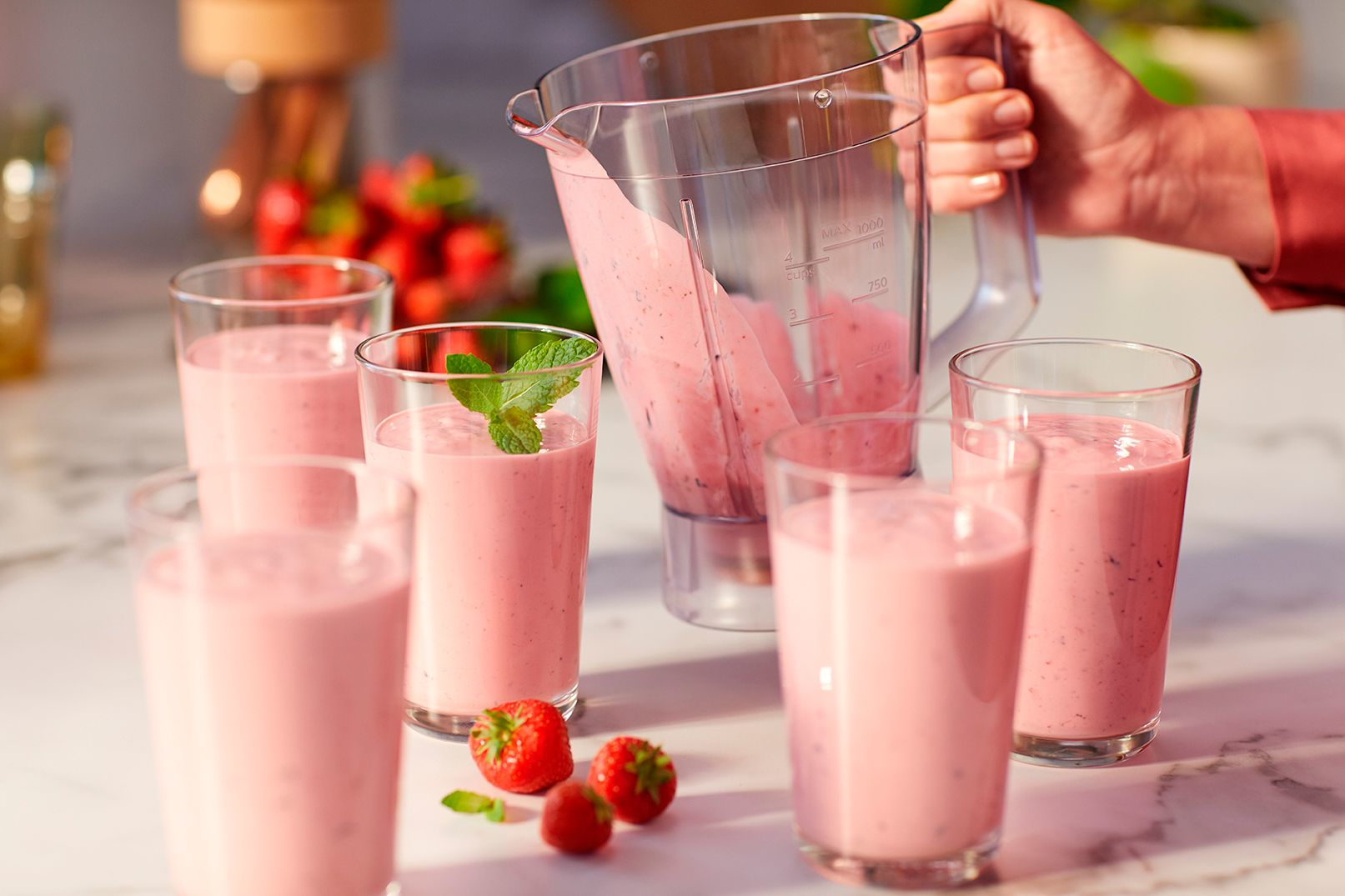 Person gießt rosa Smoothie aus einem Mixer in Gläser mit Minze. Erdbeeren auf dem Tisch.