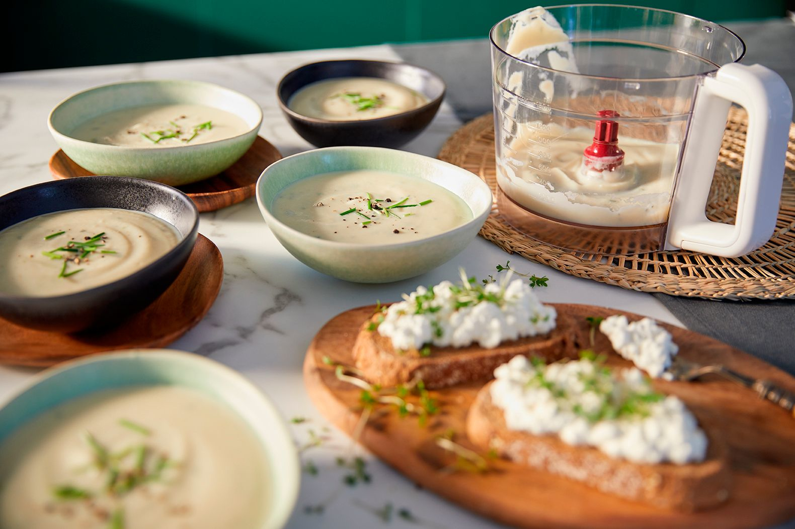 Verschiedene Schalen mit Suppe, Brot und Belägen sowie ein Foodprocessor auf einer Marmortheke.