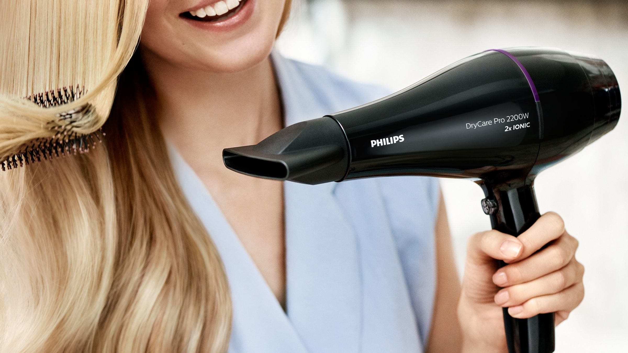 Eine Frau benutzt einen schwarzen Philips Haartrockner auf blondem Haar, lächelnd, in blauem Oberteil.