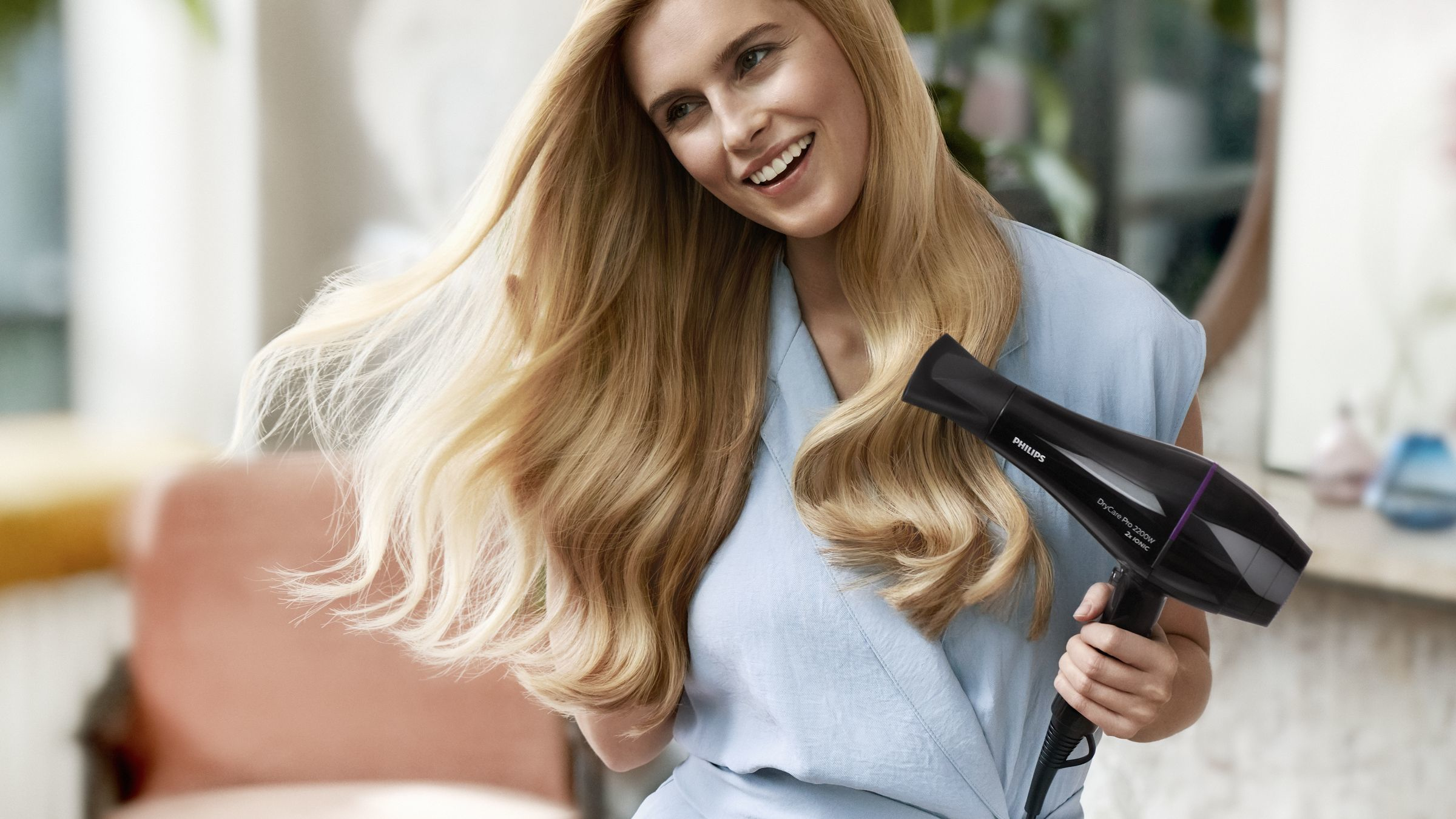 Frau lächelt, benutzt einen schwarzen Haartrockner für ihr langes blondes Haar.