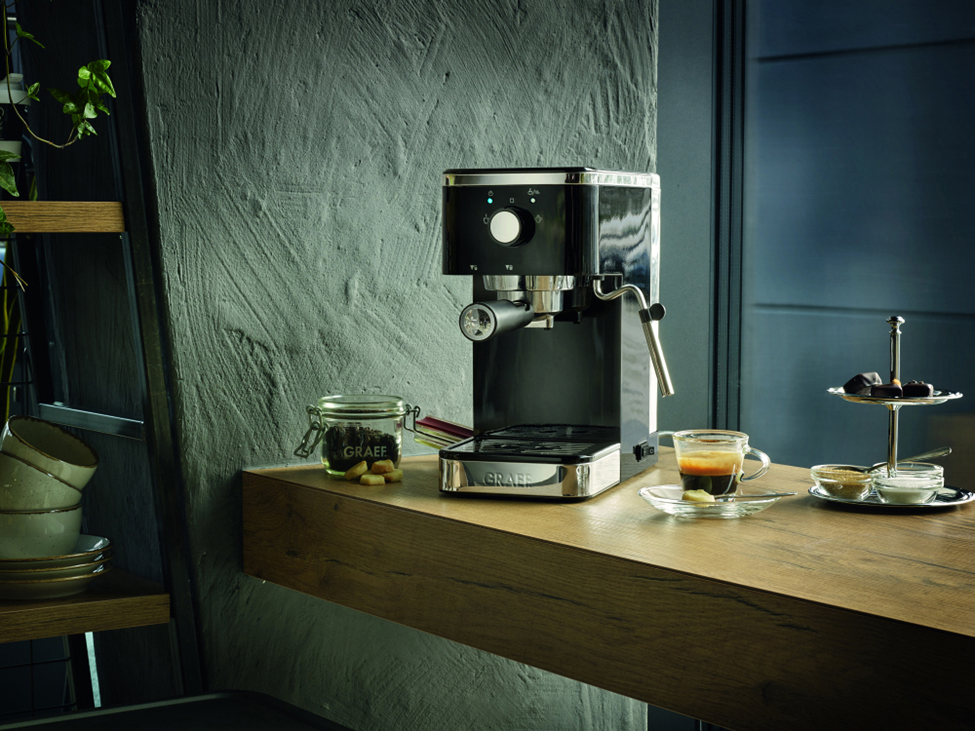 Eine Espressomaschine auf einer Holztheke mit Kaffee und Zucker sowie einem Regal mit Geschirr.