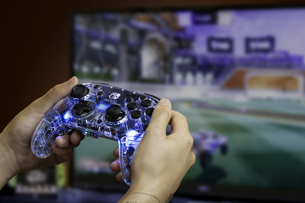 Mains tenant une manette Switch translucide avec des lumières bleues, jouant à un jeu sur une télévision.