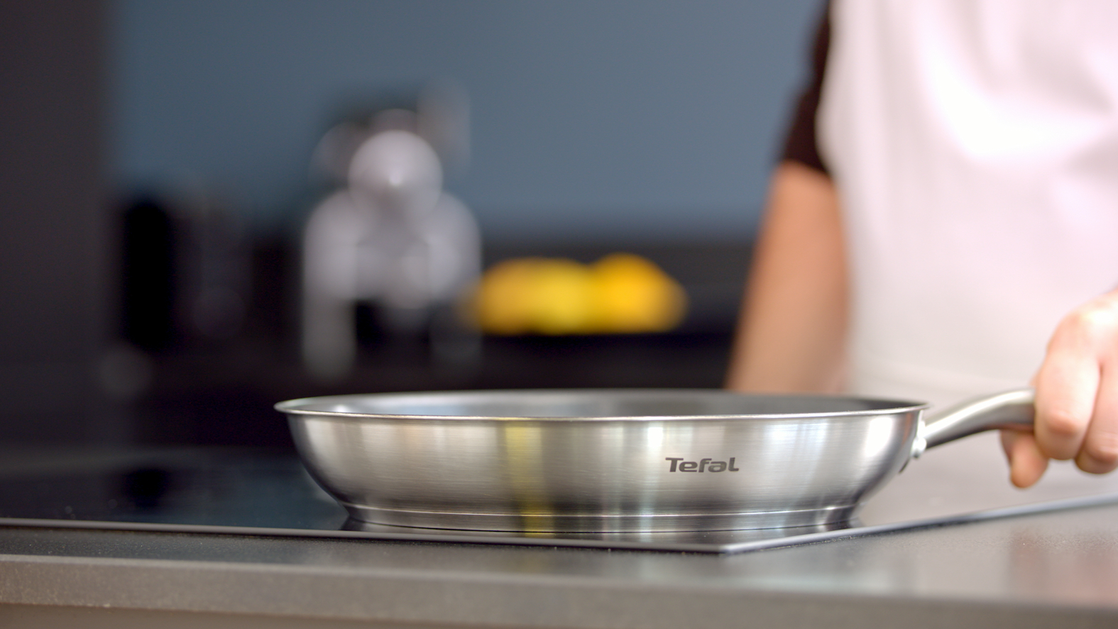 A stainless steel Tefal frying pan on a black stovetop. A hand holds the pan. The scene is in a kitchen.