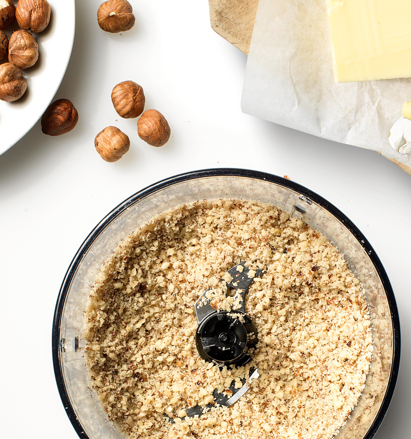 A food processor filled with finely chopped hazelnuts, plus a plate of whole nuts and butter in the background.