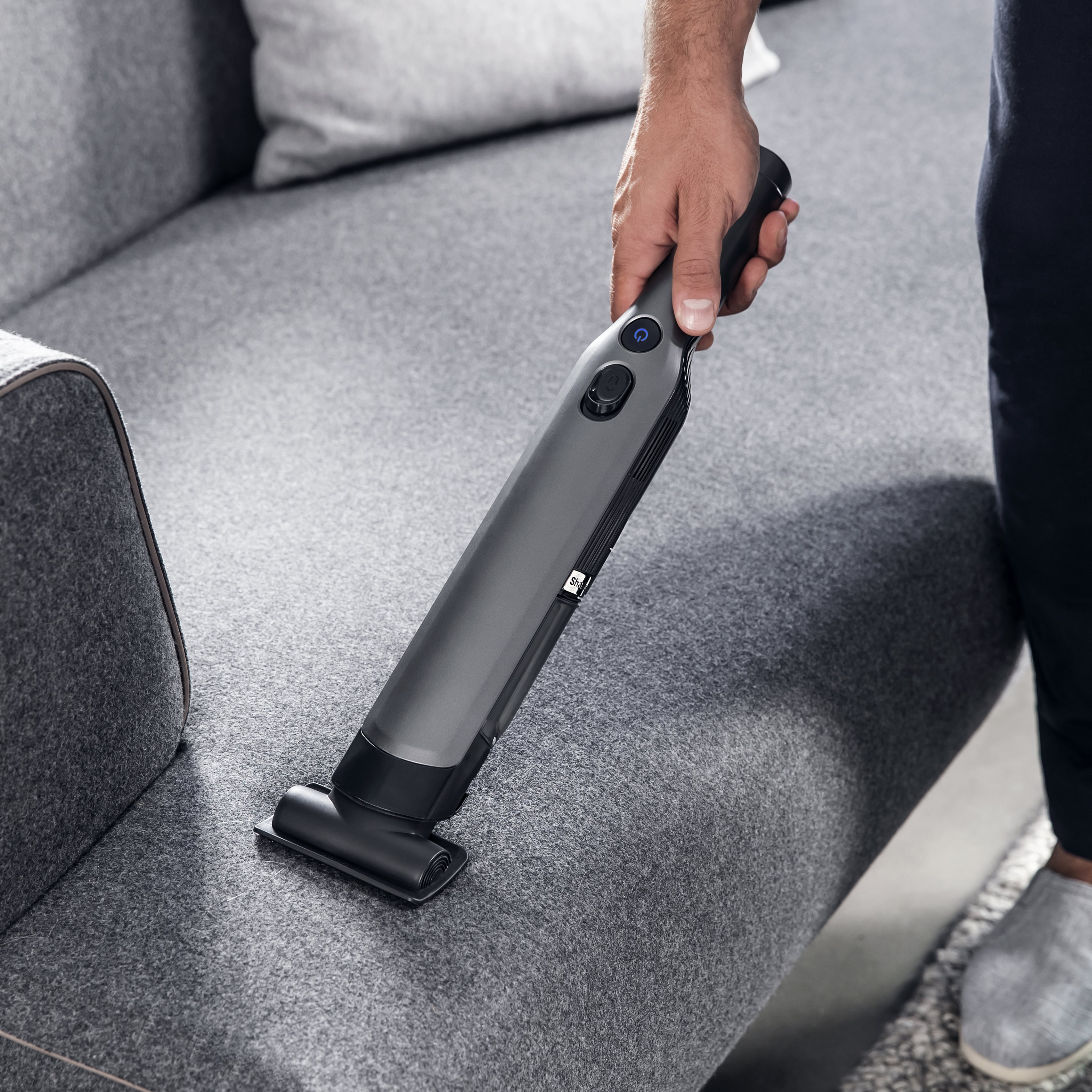 Une personne qui passe l'aspirateur sur un canapé gris avec un aspirateur à main.