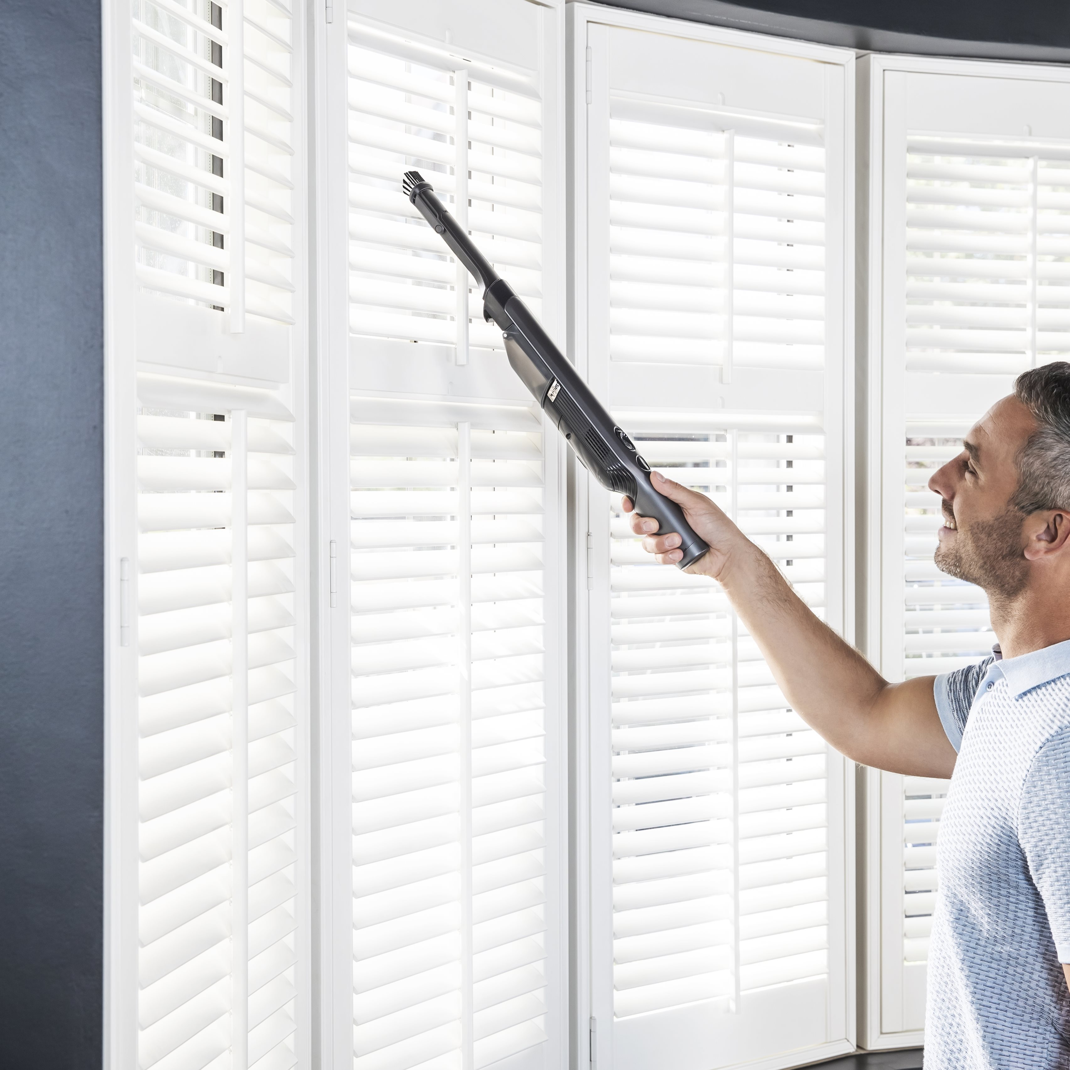 Un homme nettoyant des volets blancs avec un aspirateur à main. Il regarde les volets.