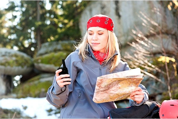 Woman in nature holds map and radio. She wears a red bandana, with green trees and rocks.