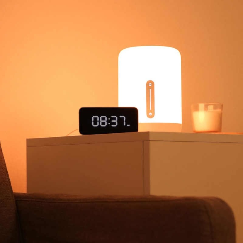 Una mesita de noche moderna con un reloj digital, una lámpara y una vela, con un brillo anaranjado.