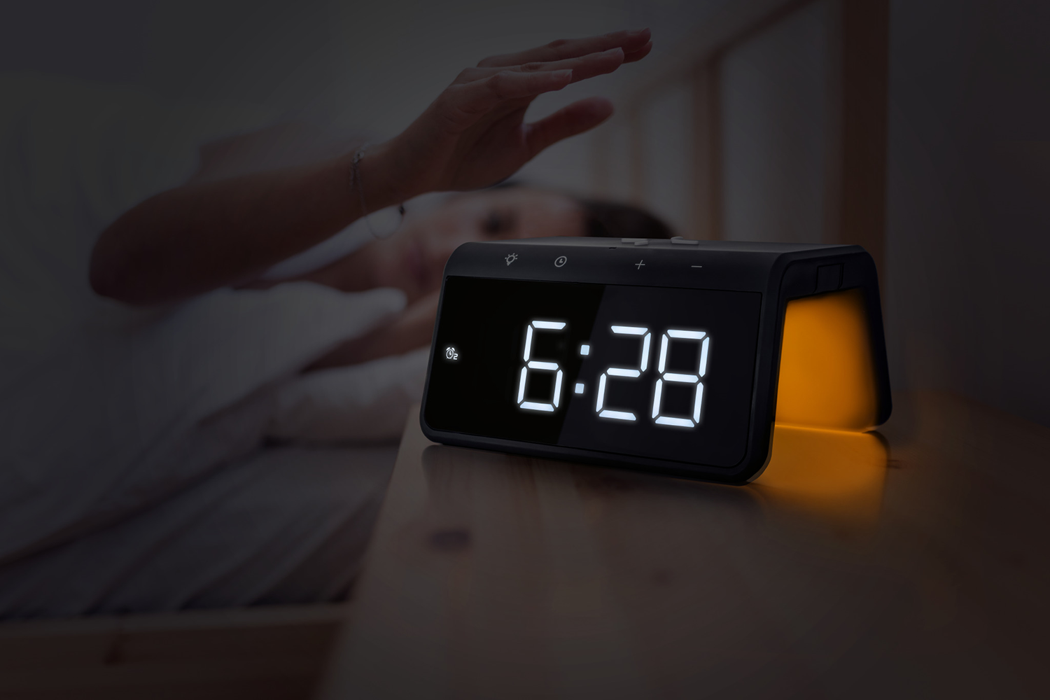 An alarm clock displaying 6:28 on a nightstand. A person's hand reaches towards it, in a dark room.
