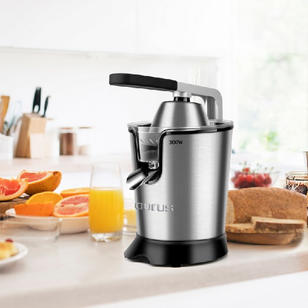 Exprimidor en una encimera con fruta y zumo. Electrodoméstico plateado y negro. Ambiente de cocina.