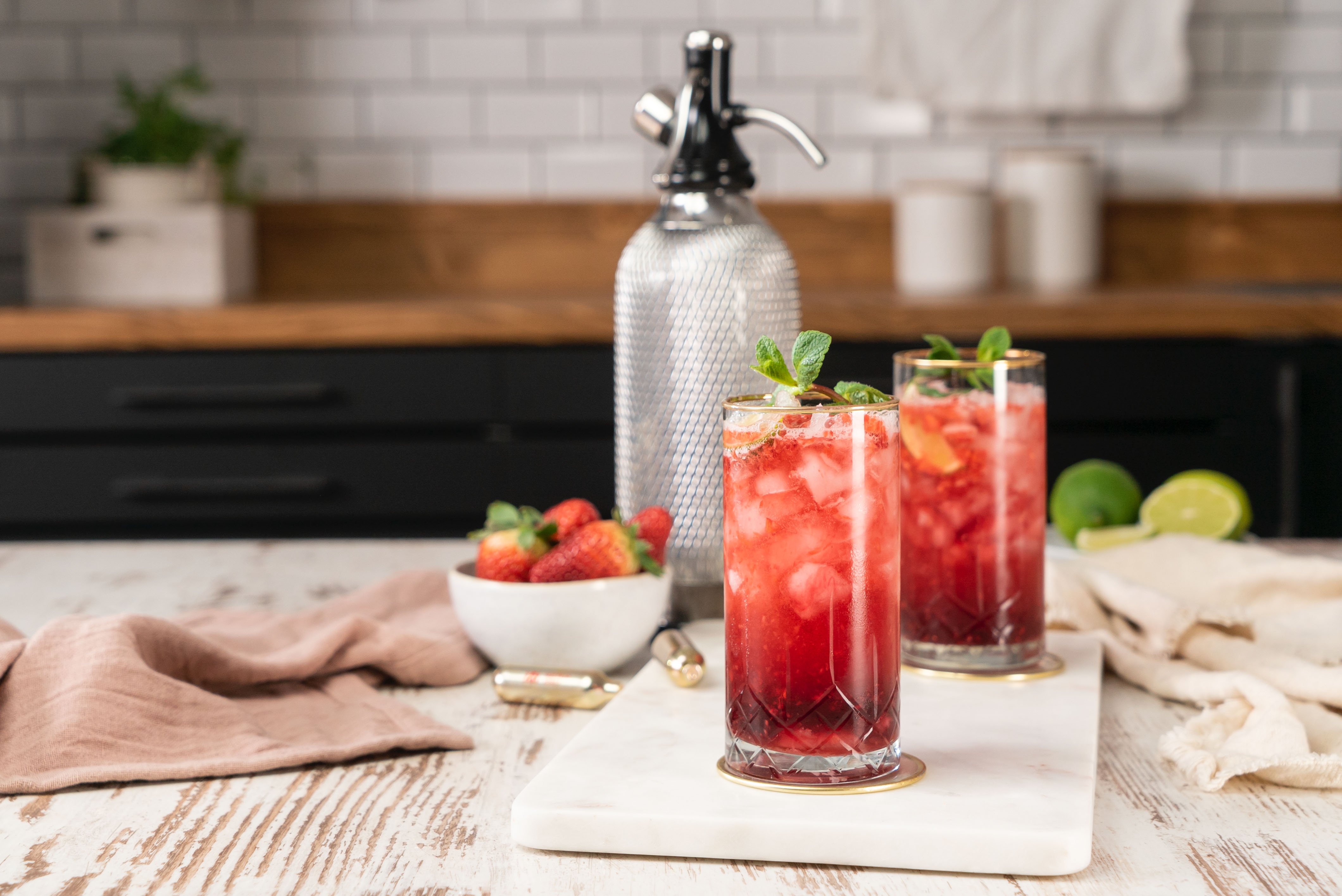 Zwei Erdbeer-Cocktails mit Minze, eine Soda-Siphon und eine Schüssel Erdbeeren auf Marmorplatte.