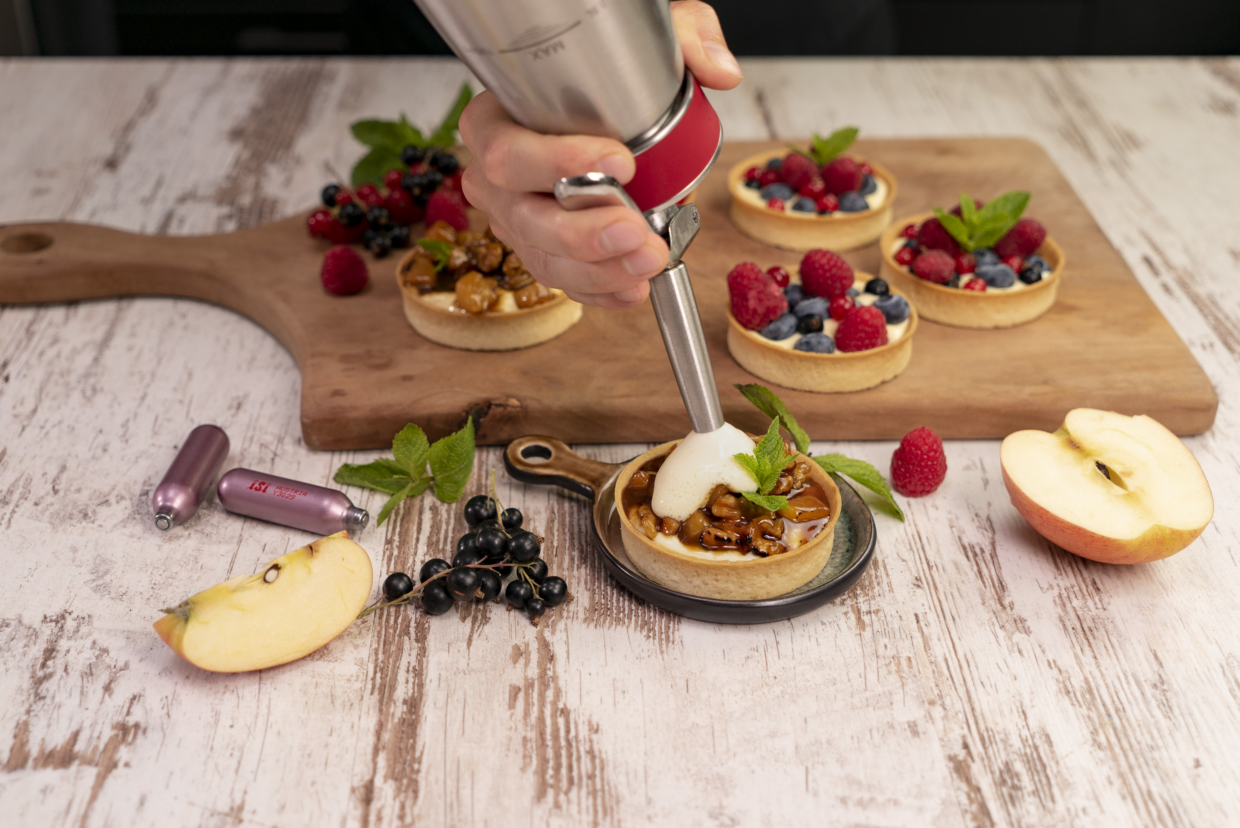 Eine Person dekoriert Törtchen mit Schlagsahne mit einem Spender. Beeren, Apfel und Minze.