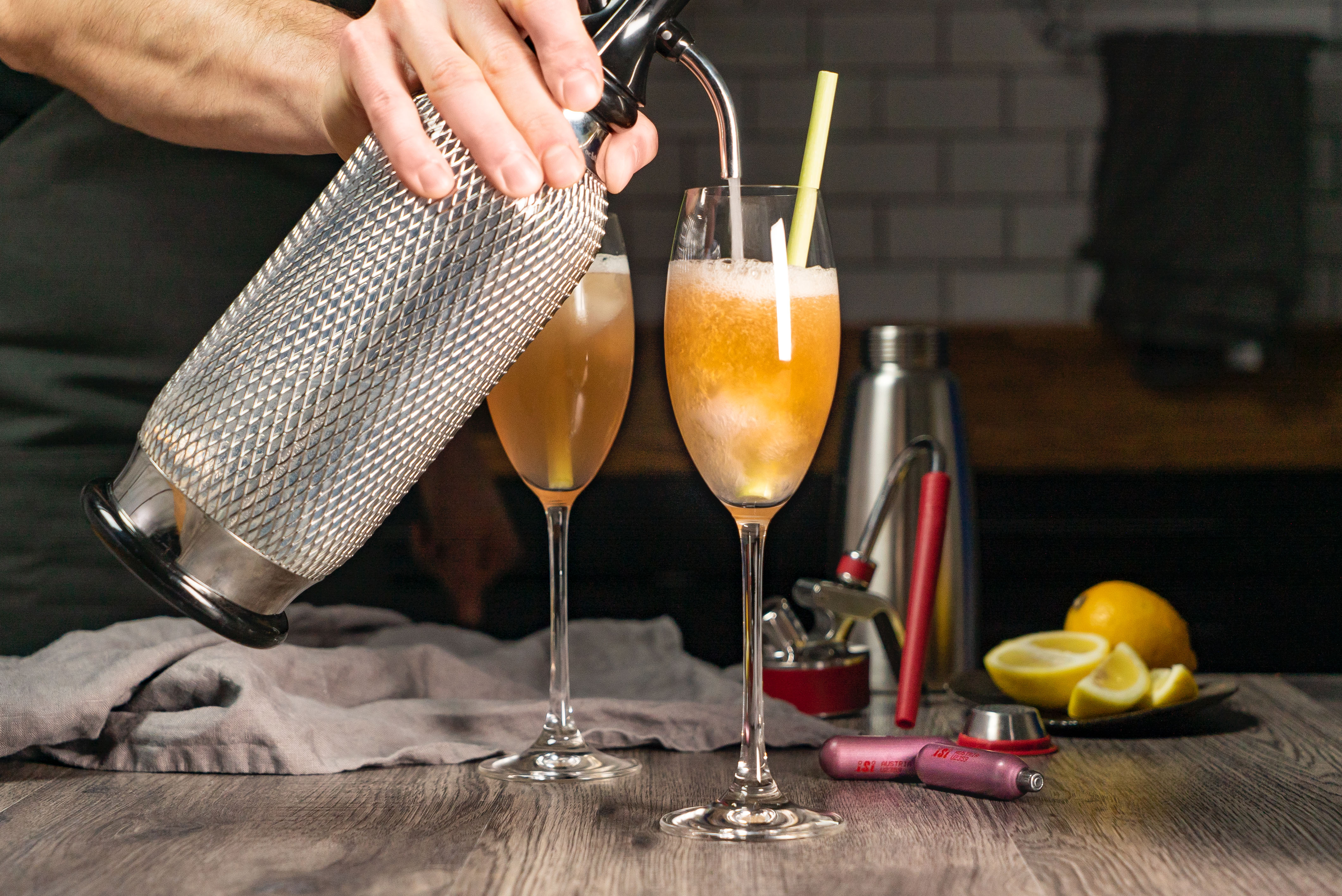 Ein Barkeeper gießt einen Cocktail mit Strohhalm in ein Glas. Ein weiteres Glas daneben.