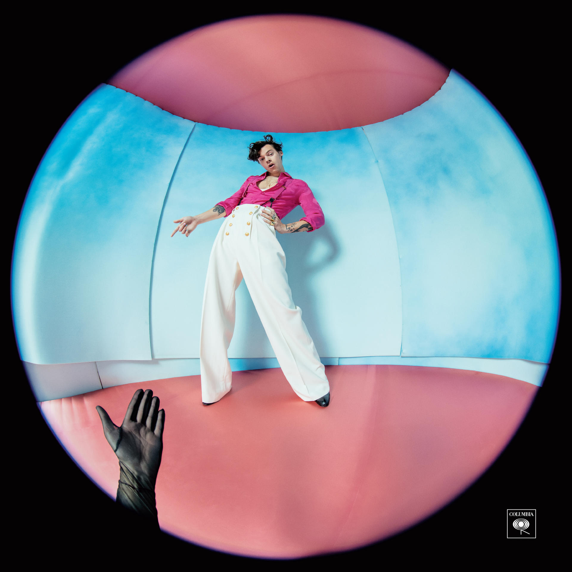 Harry Styles in a pink shirt and white pants, standing in a pink and blue studio.