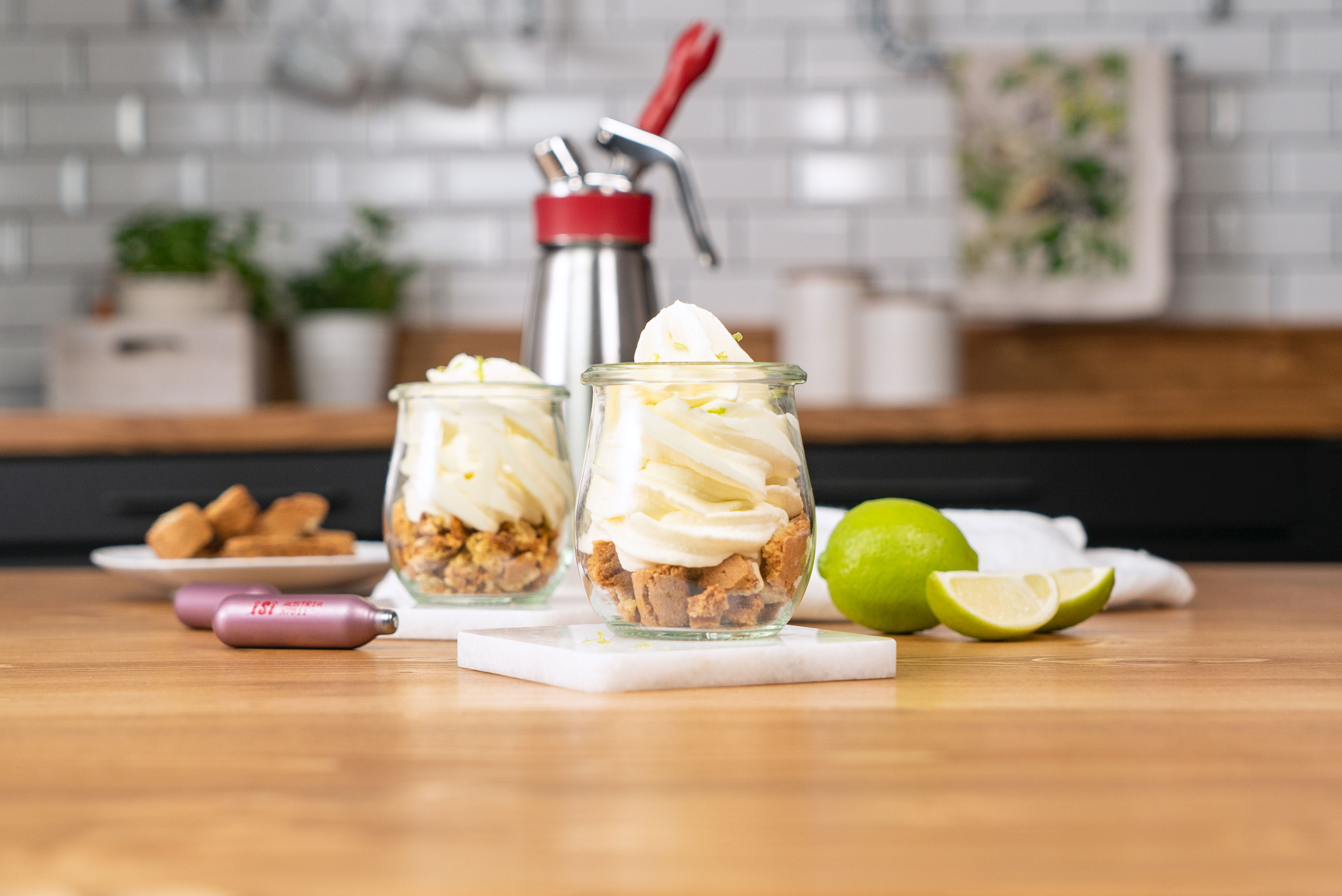 Zwei Gläser mit Dessert, Schlagsahne obenauf; Limettenscheiben, Kekse und Sahnespender daneben.