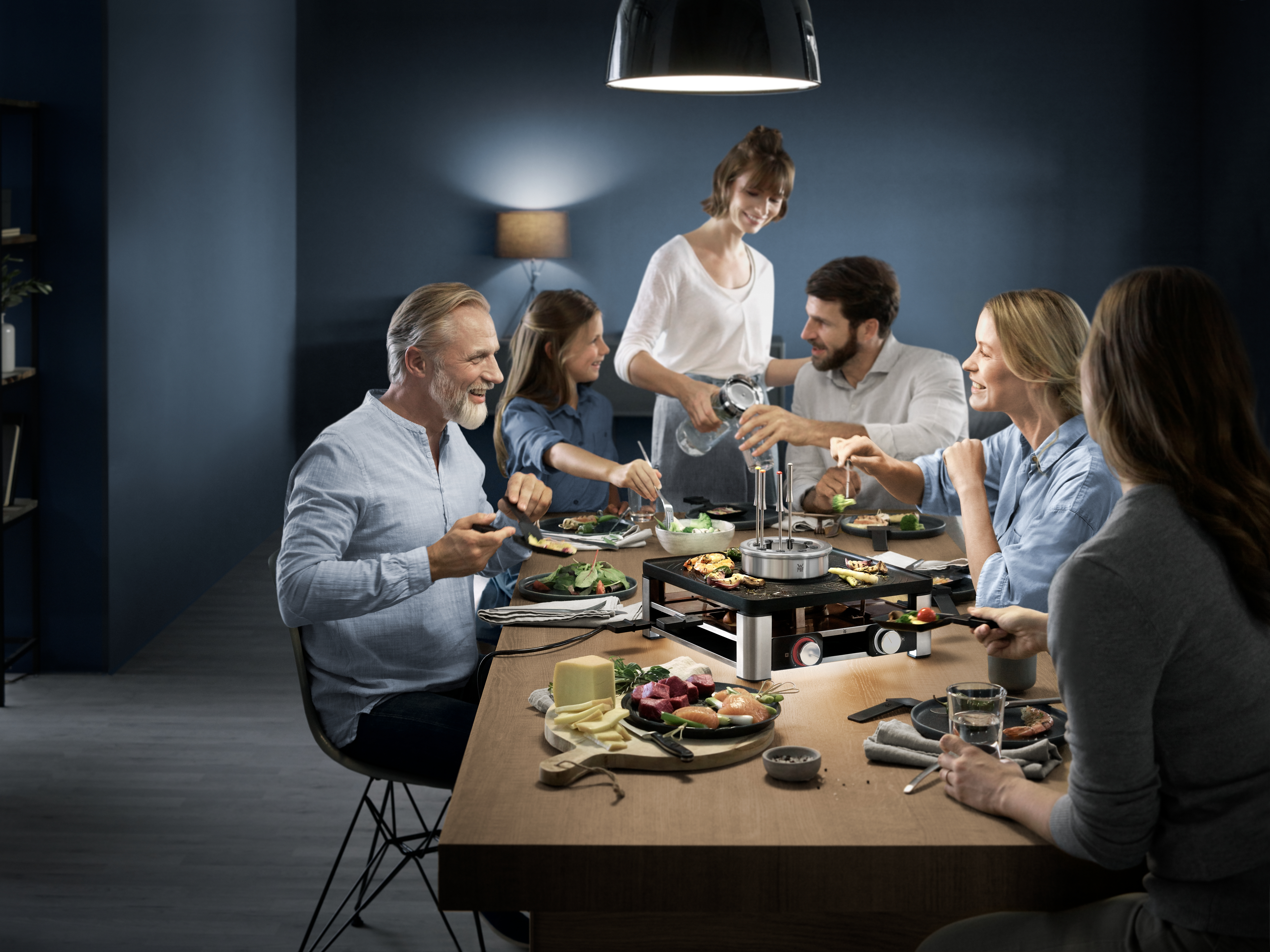 Eine Familie genießt eine Mahlzeit an einem Tisch mit einem Raclette-Grill in gemütlicher Atmosphäre.