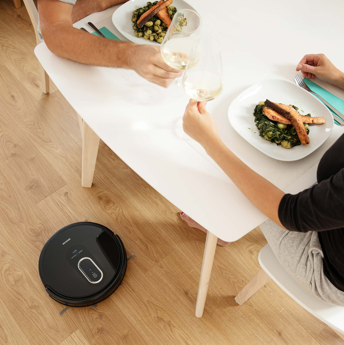 Egy pár koccint borral, ételtányérokkal és egy fekete robotporszívóval.