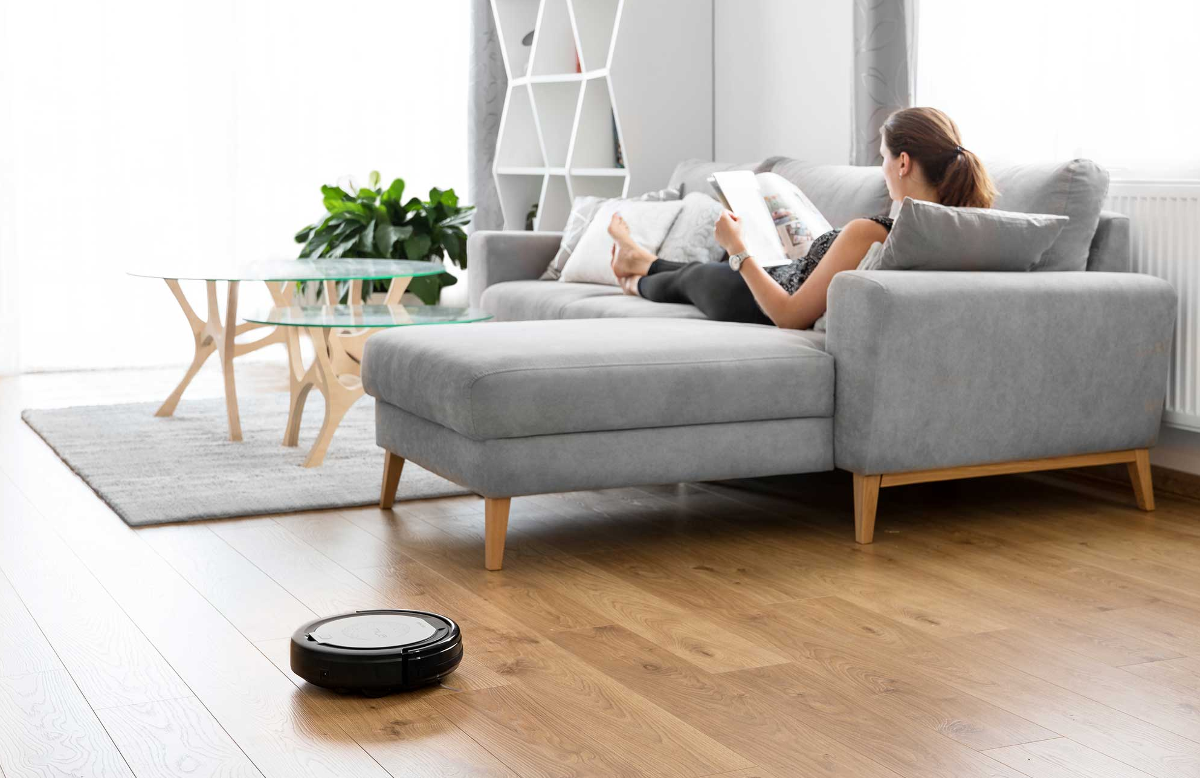 Egy nő egy szürke kanapén olvas egy könyvet, egy robotporszívó a padlón. Egy asztal és egy növény a helyszínen.