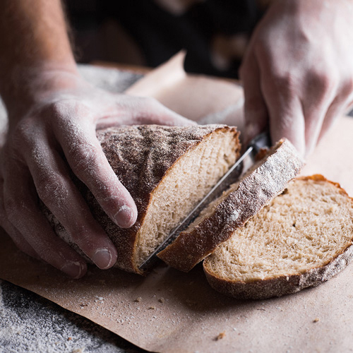 Hände schneiden Brot mit einem Messer auf einem Holzbrett, etwas Mehl ist verstreut.