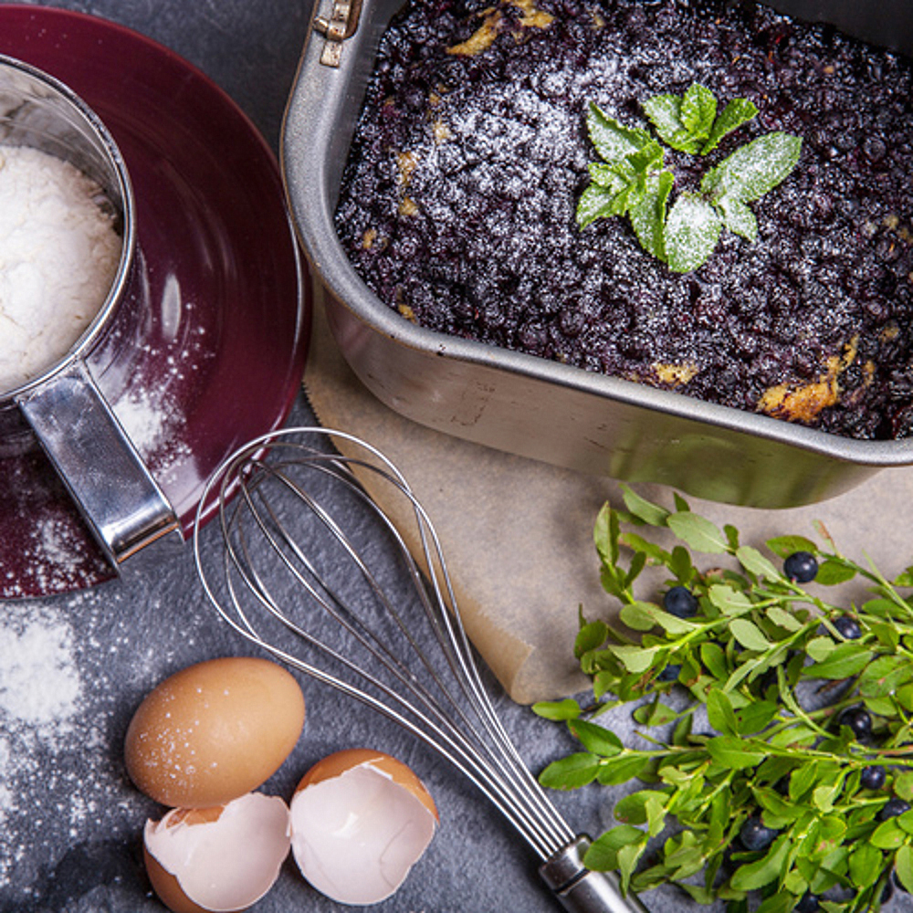 Kuchen in einer Metallpfanne mit Puderzucker, Minze und Blaubeeren auf dunkler Oberfläche mit Zutaten.