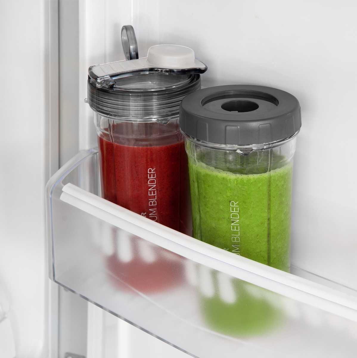 Két smoothie-palack a hűtőben. Egy piros, egy zöld, mindkettő fekete fedéllel. Átlátszó polcon.