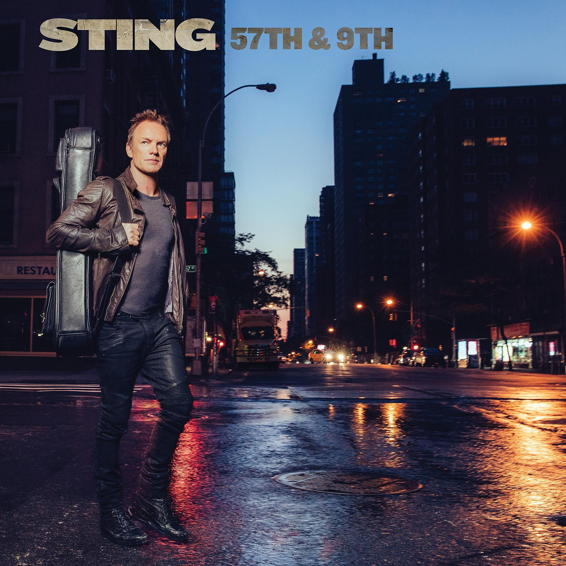 Sting auf einer Straße in der Dämmerung, mit Gitarrenkoffer. Gebäude und Straßenlaternen im Hintergrund.