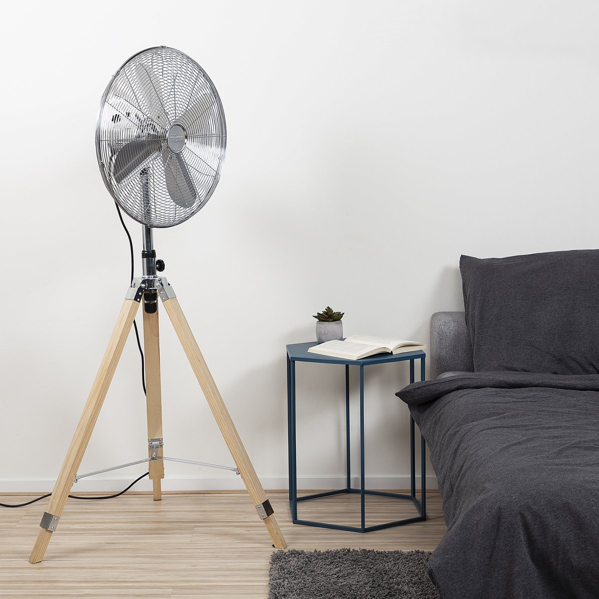 Silberner Ventilator auf Holzstativ, neben kleinem blauen Tisch und Bett mit grauen Laken.
