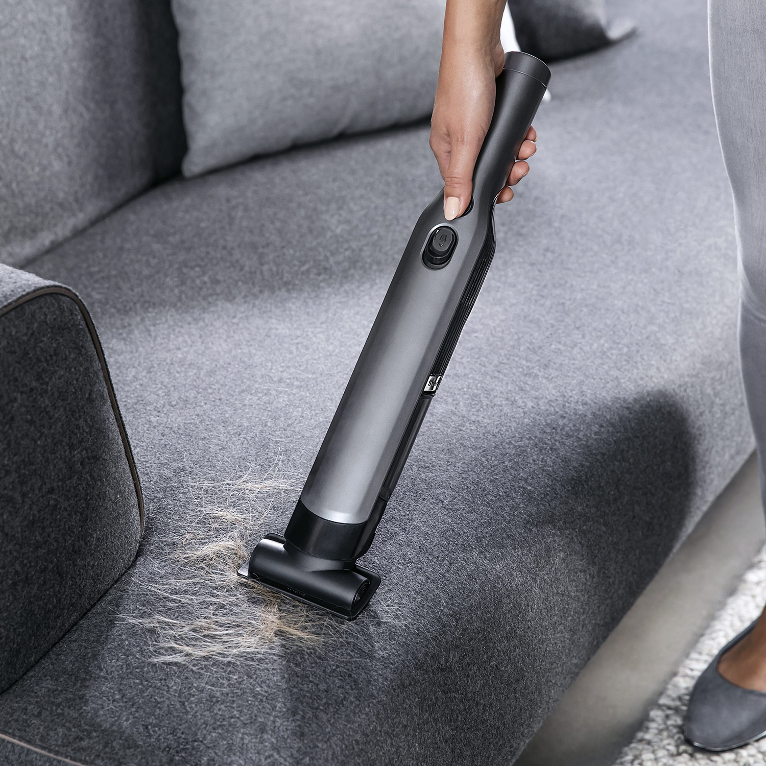 A person vacuums a gray sofa with a handheld vacuum cleaner. The sofa has pet hair on it.